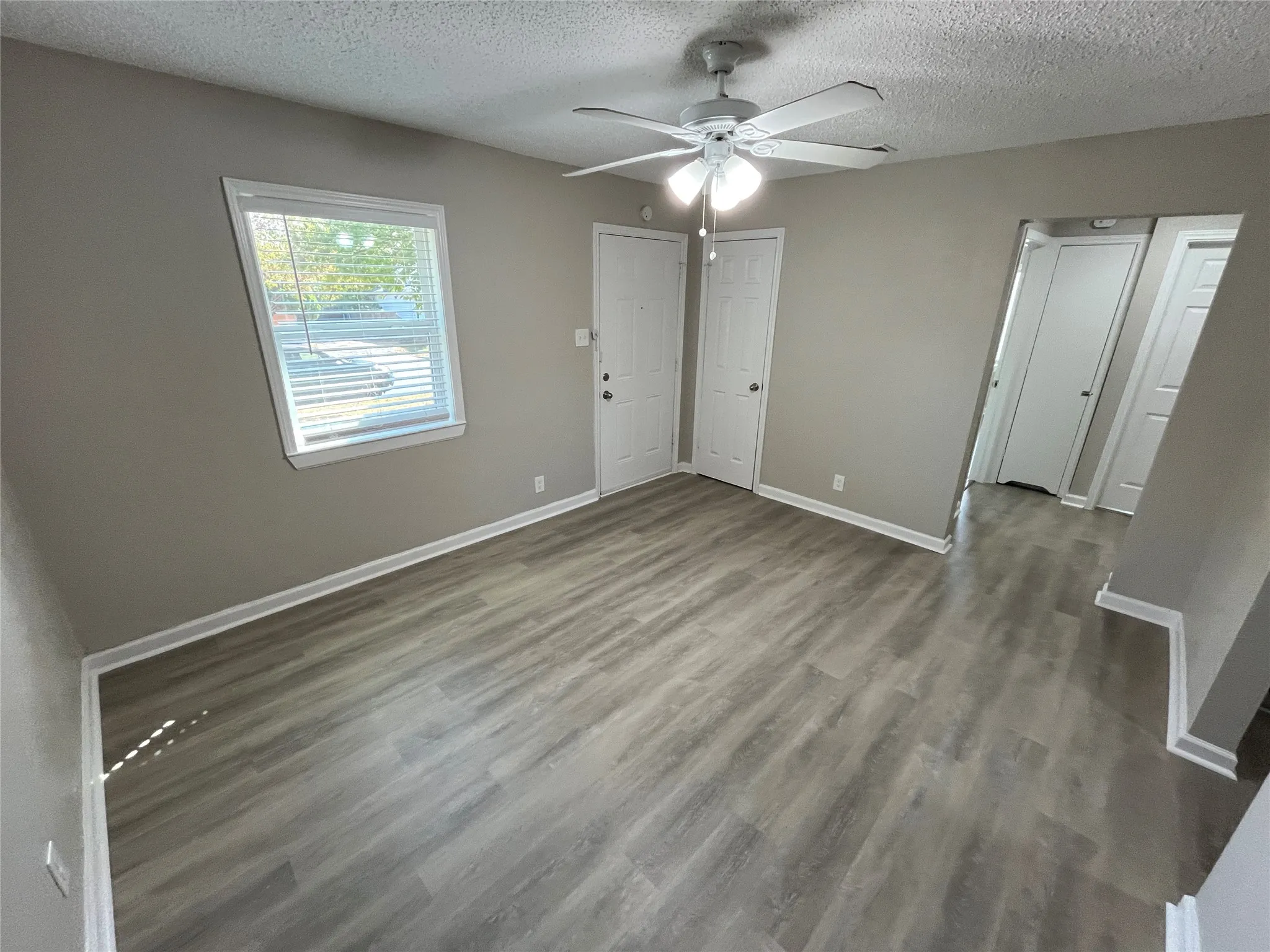 Gray wood-finish flooring, white ceiling fan with integrated lighting, window with blinds, and neutral wall paint