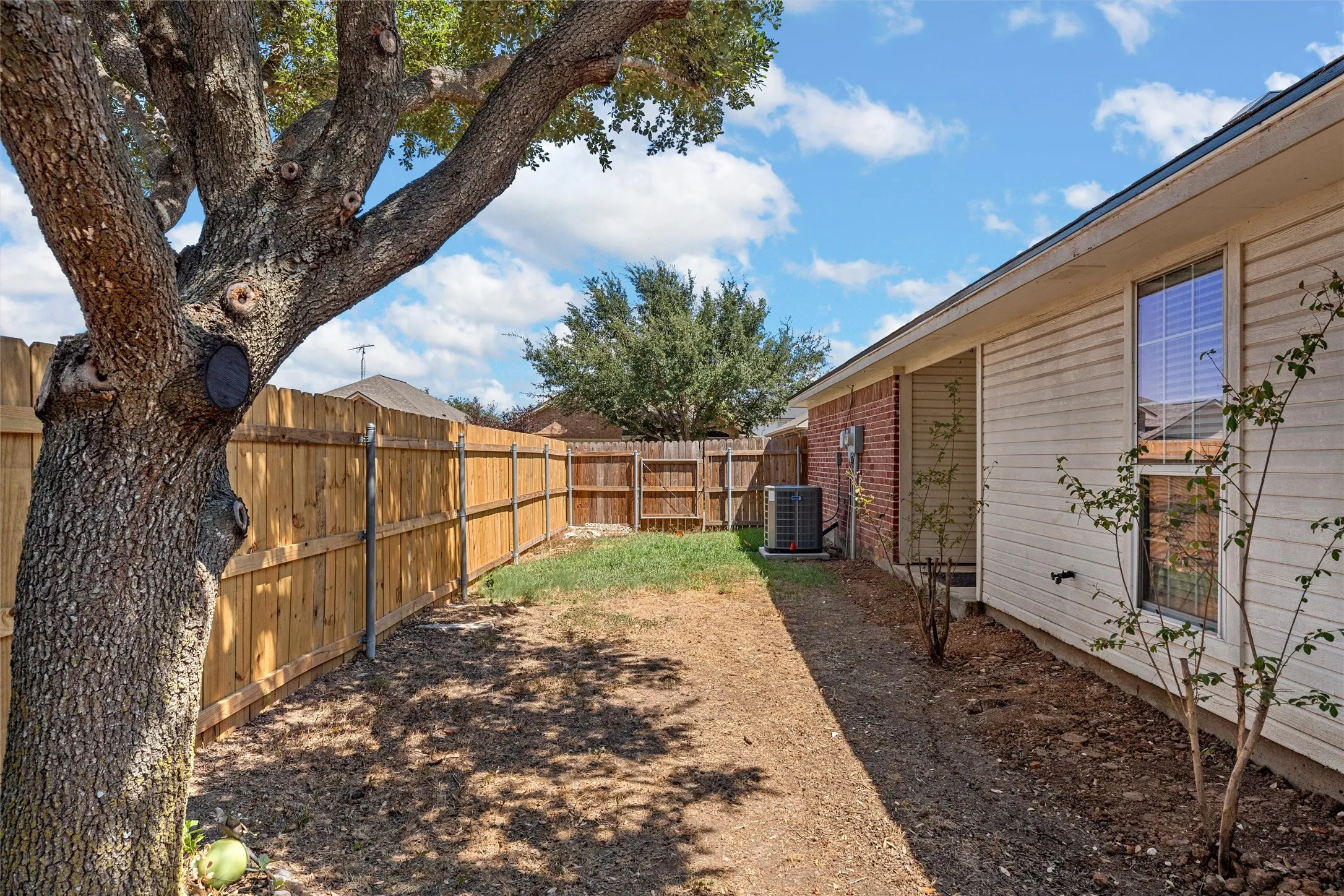 Fully fenced backyard featuring a large mature tree, established shrubbery, and brick and siding exterior