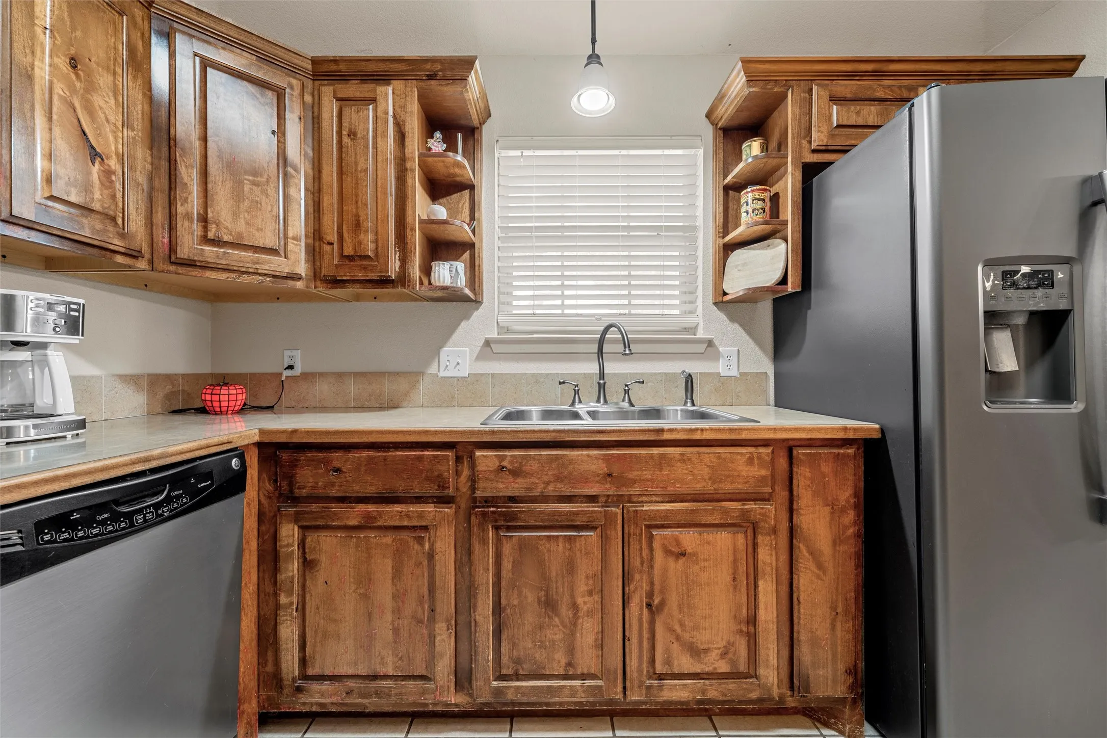 Wood-finish cabinetry with open shelving, stainless steel double basin sink with a chrome faucet, light-toned countertop, and tile backsplash