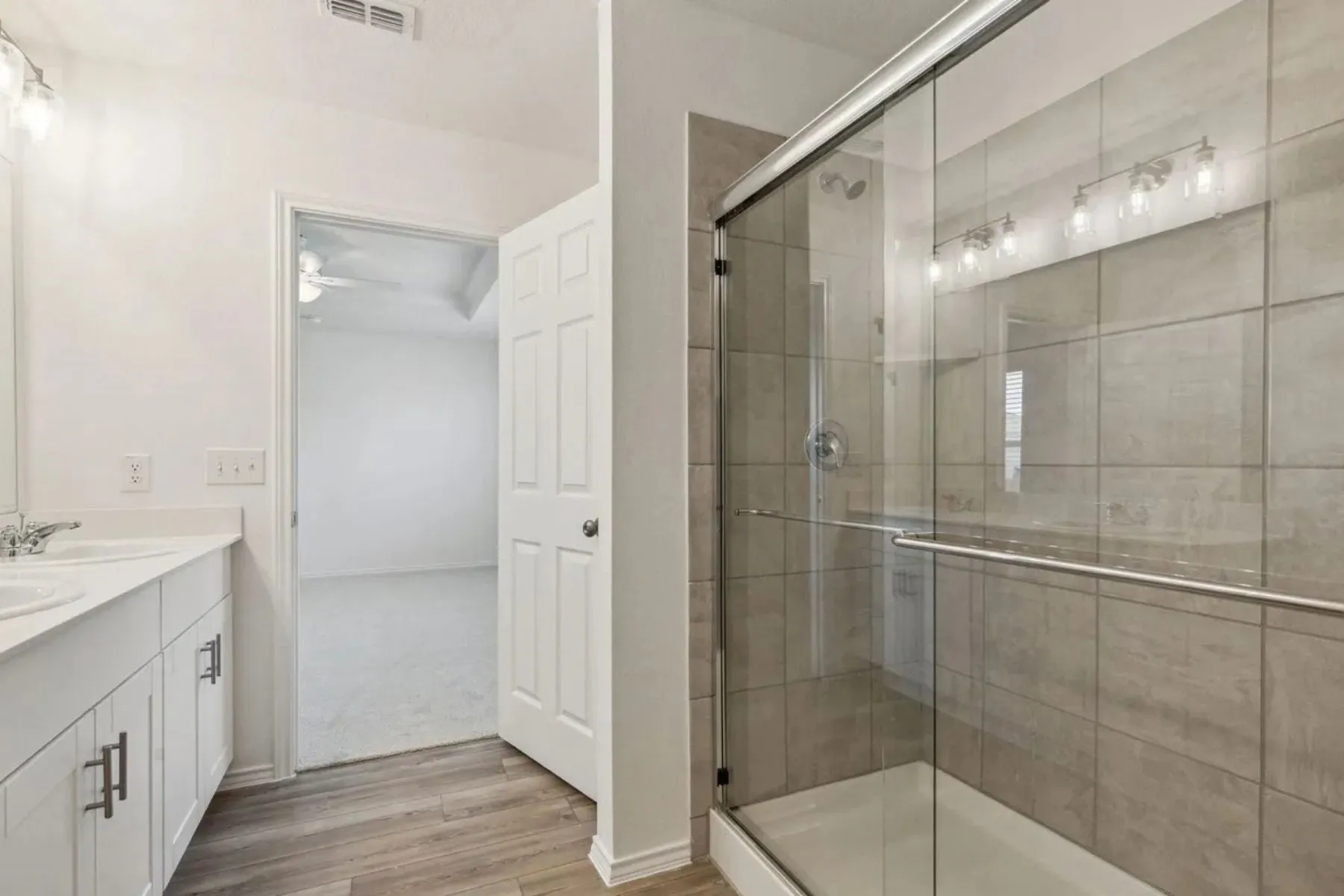 Dual vanity with white cabinetry, wood-finish flooring, and a glass-enclosed shower with tile surround