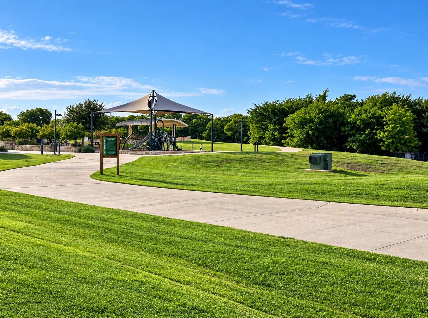 Paved walking path, manicured green lawn, mature trees, and a shaded play structure