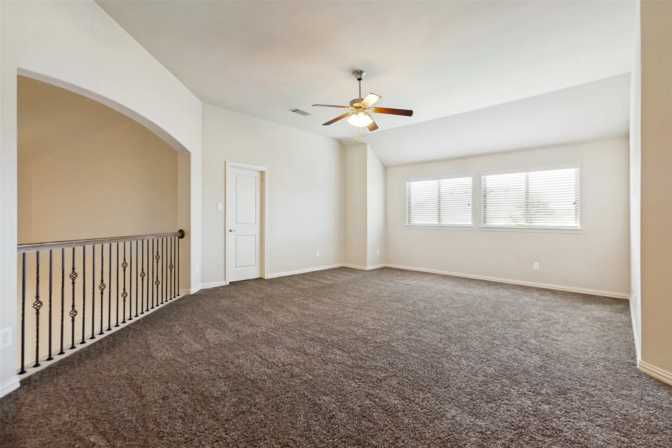 Spacious carpeted room featuring a ceiling fan, multiple windows with blinds, and an arched opening with an iron railing