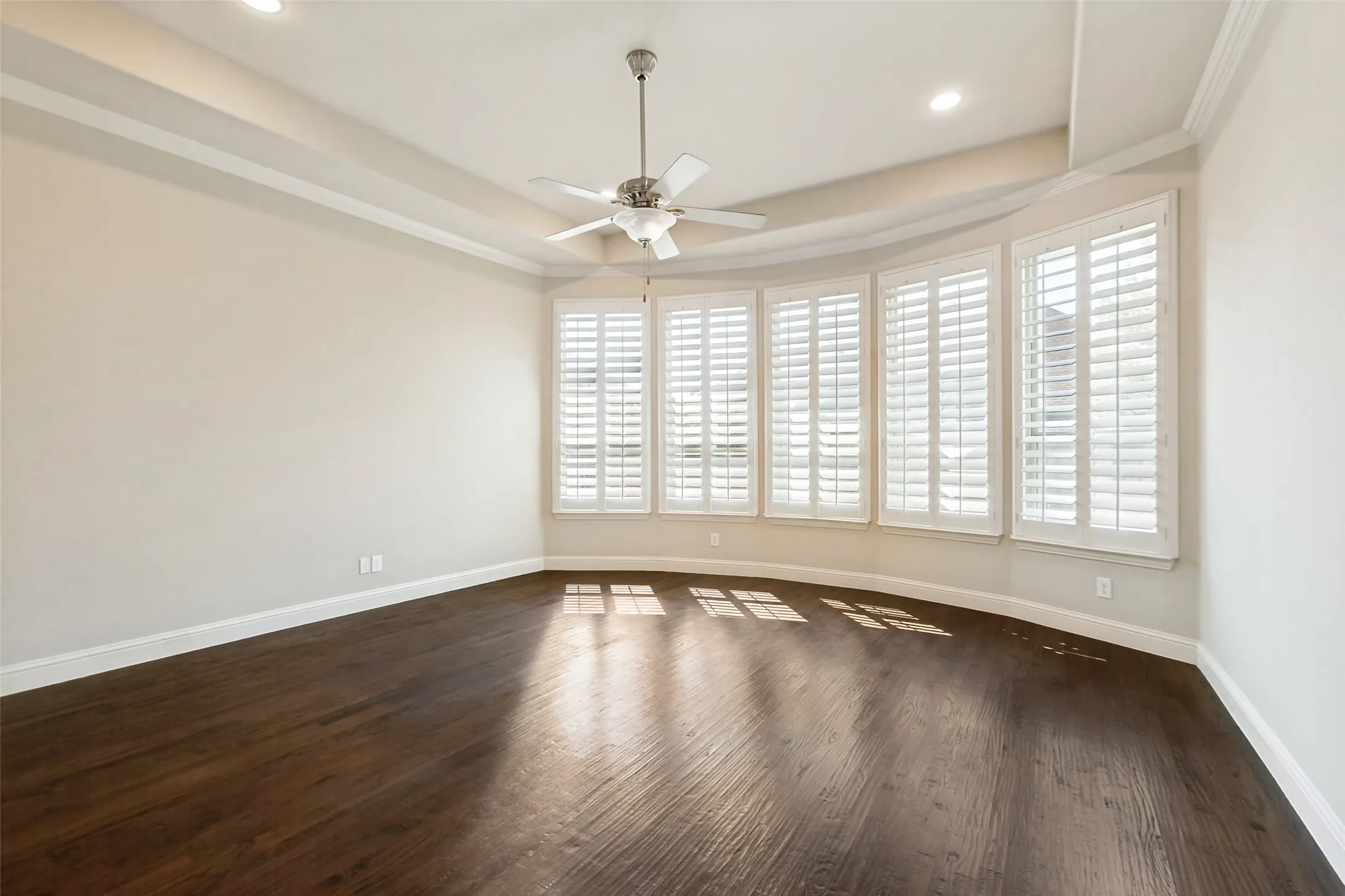 Tray ceiling with recessed lighting, ceiling fan, wood-finish flooring, and bay windows with plantation shutters