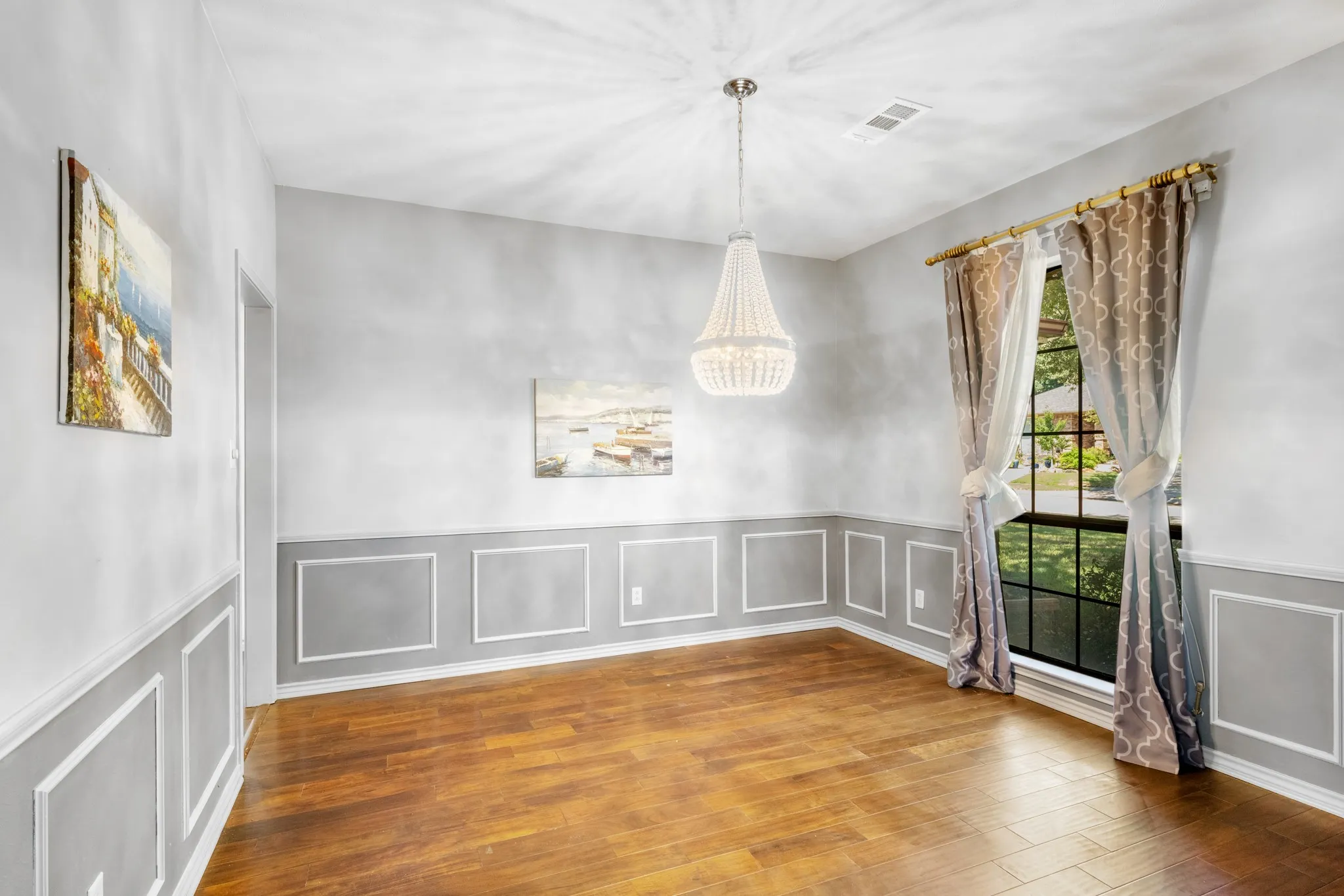 Elegant formal dining room featuring rich wood flooring, sophisticated decorative wall molding, and soft neutral tones. A stunning crystal chandelier adds a touch of glamour, while oversized windows provide abundant natural light and picturesque views of the beautifully landscaped surroundings.