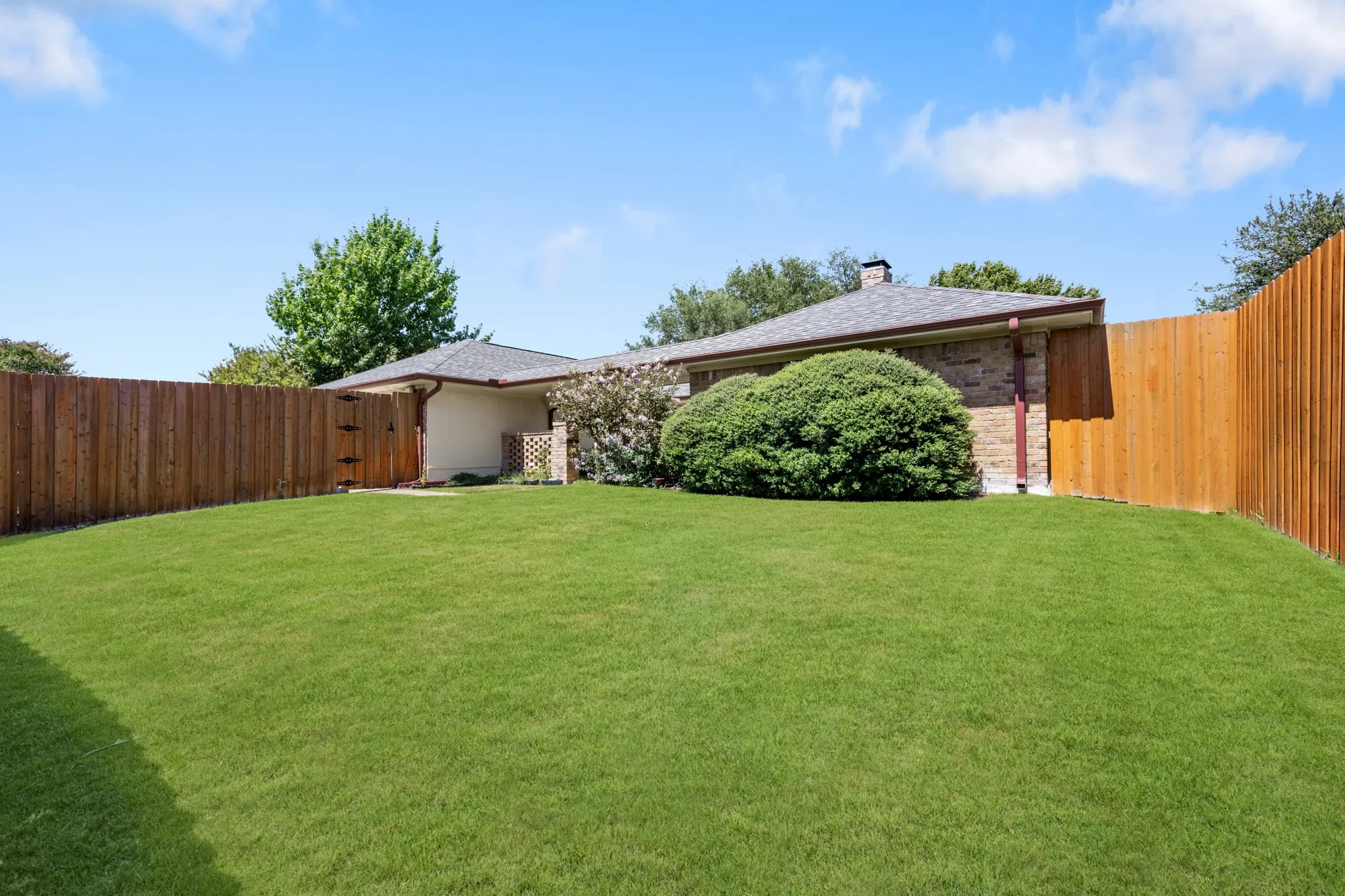 Beautifully landscaped backyard offering an expansive, lush lawn framed by mature greenery and a privacy-enhancing wood fence. A covered patio provides a comfortable outdoor retreat, creating a wonderful setting for relaxing, entertaining, and enjoying the outdoors. *Grass enhanced to show potential*