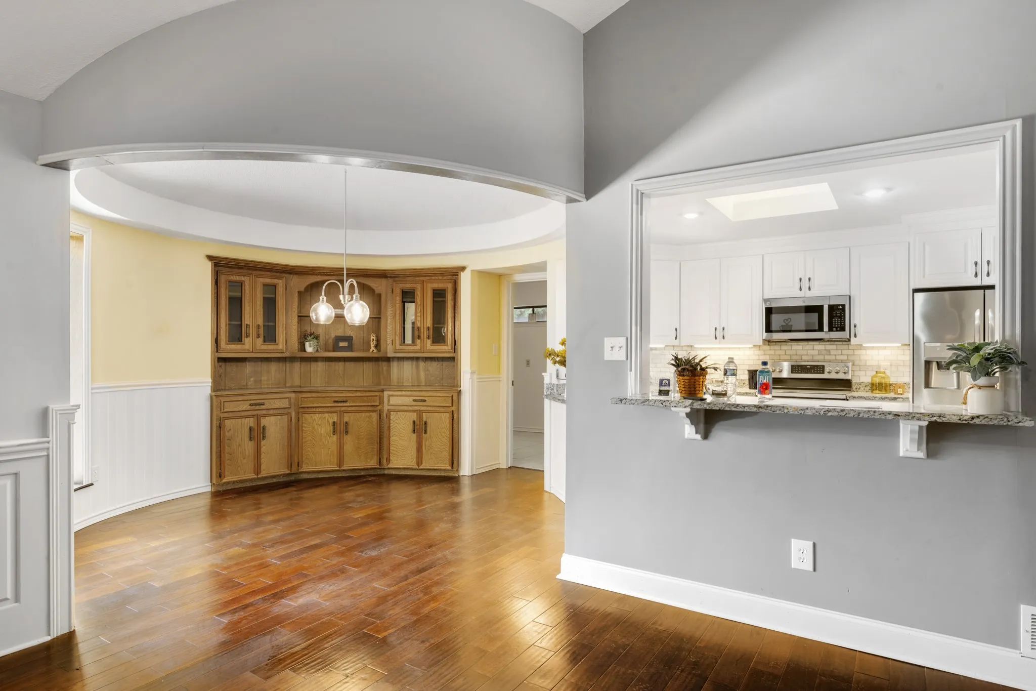 Charming breakfast nook featuring a curved architectural design, rich wood flooring, classic wainscoting, and a beautiful built-in wood hutch with glass-front cabinetry. Elegant pendant lighting and abundant natural light create a warm, inviting space for casual dining and morning gatherings.