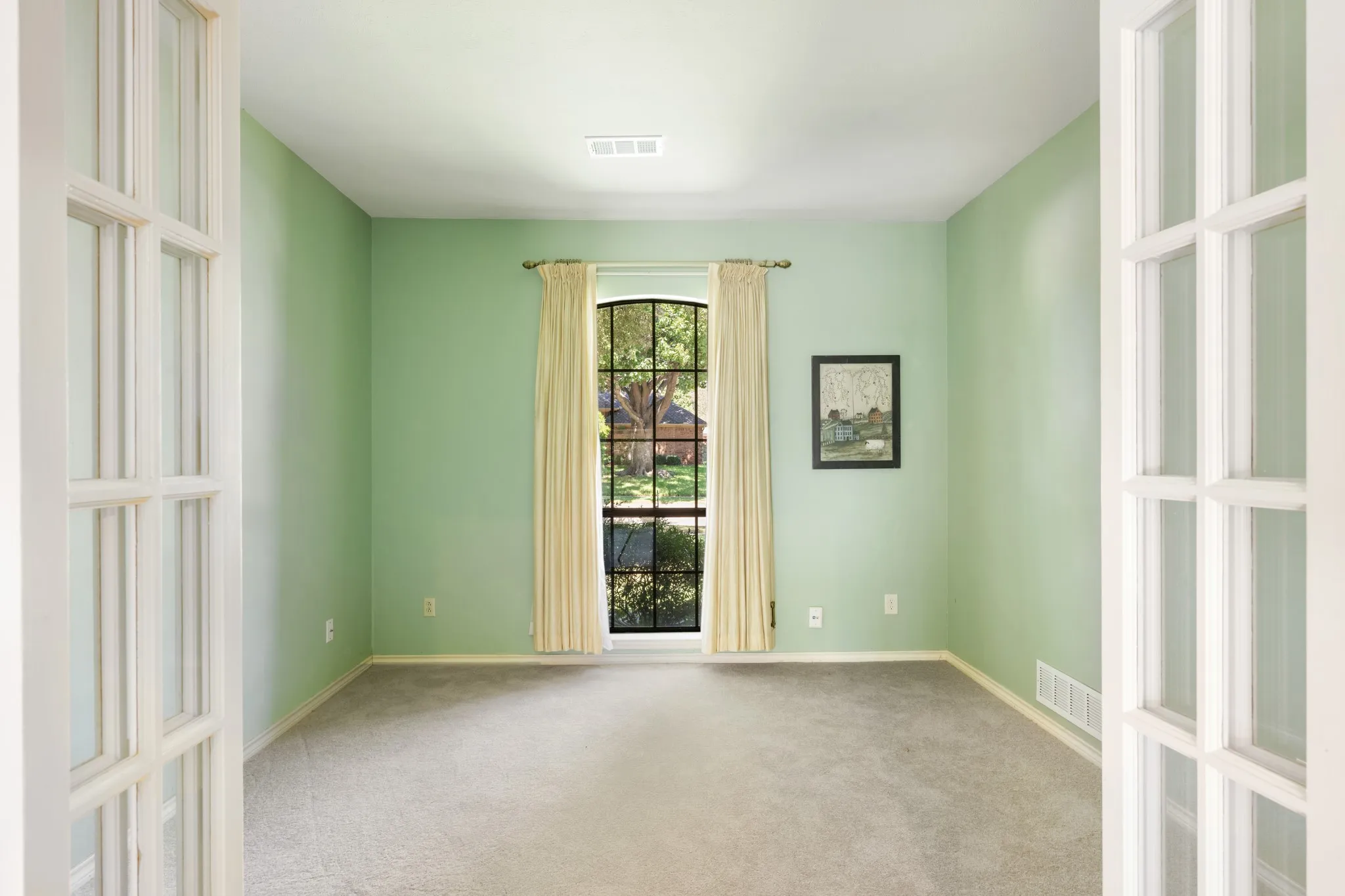 Light-filled private office featuring French doors, soft green walls, plush carpeting, and a charming arched window overlooking the front yard. The inviting space offers a peaceful setting for a home office, study, or flex space.