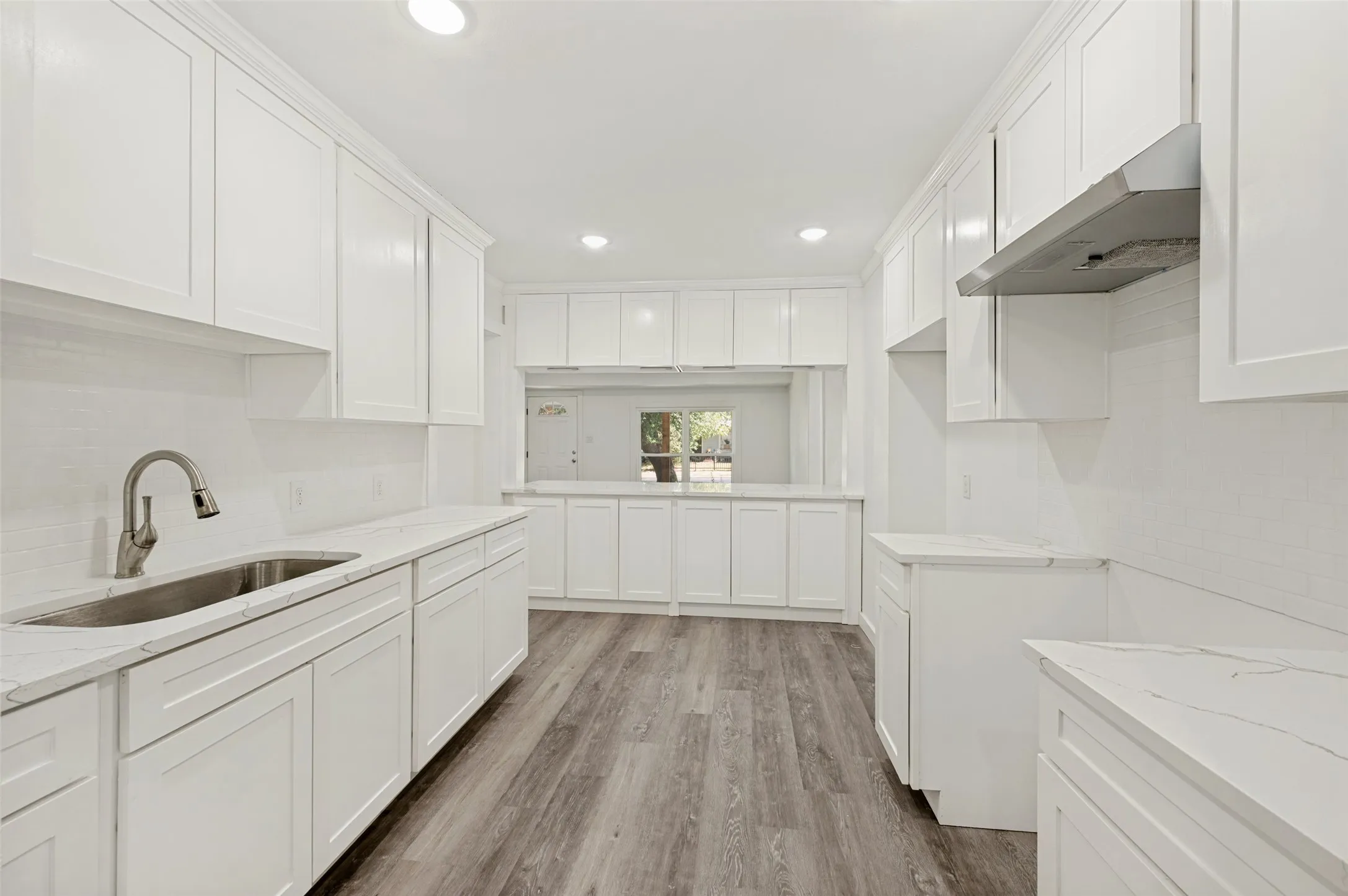 White shaker cabinetry, light-toned countertops, stainless steel sink with gooseneck faucet, horizontal tile backsplash, and wood-finish flooring
