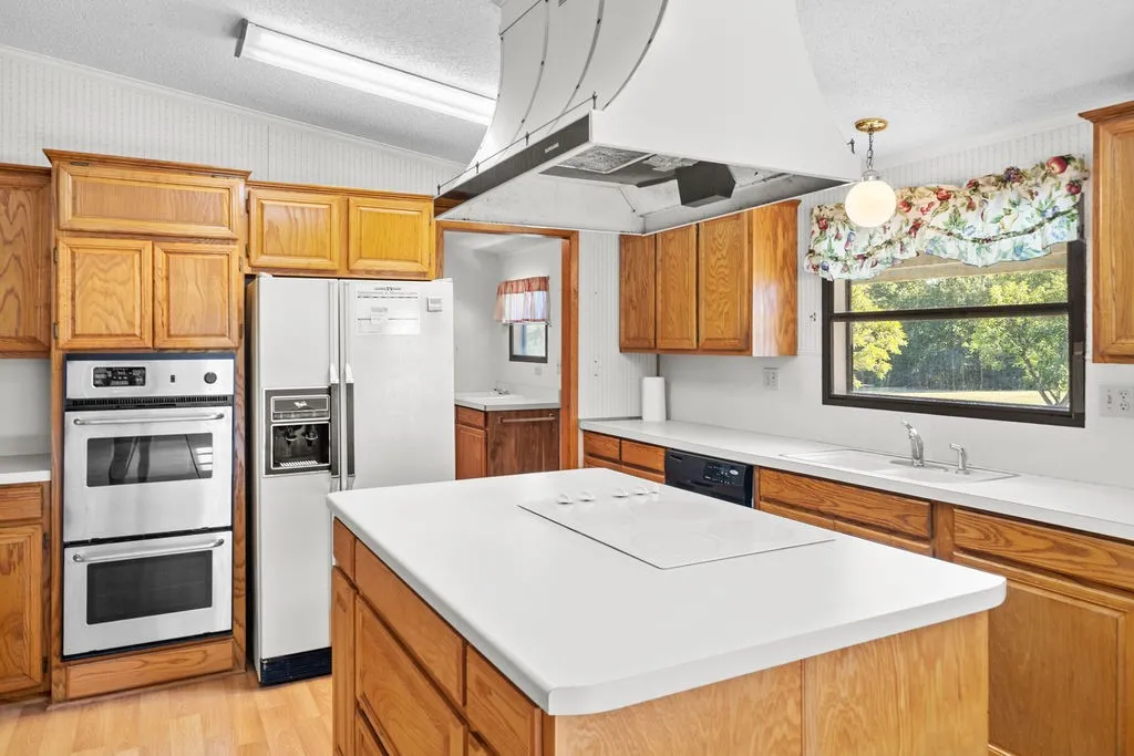 Kitchen featuring wood cabinetry, white countertops, a center island with a cooktop, a double wall oven, and a stainless steel refrigerator