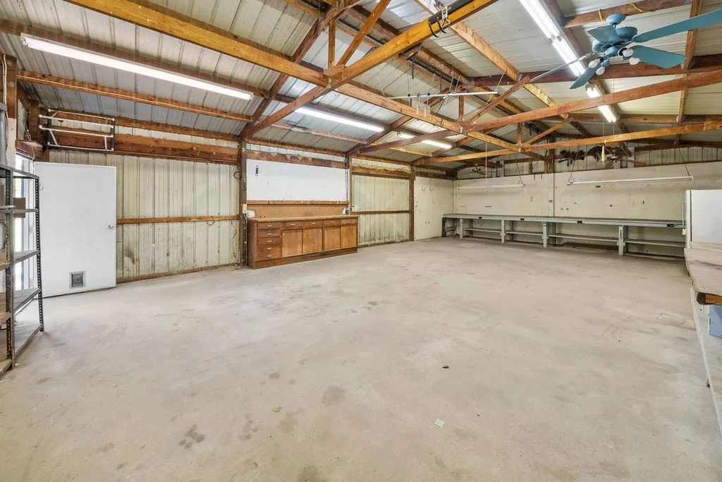 Spacious utility building with exposed wood beams, concrete flooring, fluorescent lighting, and built-in workbench areas