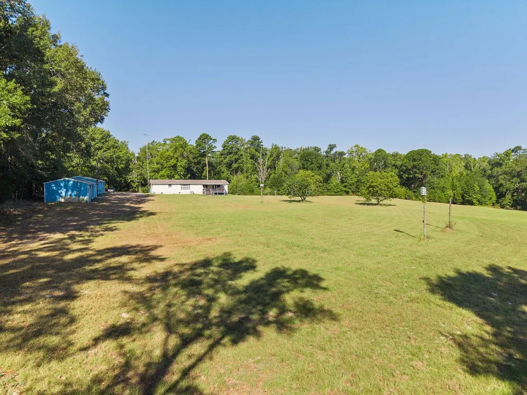 Expansive green field featuring scattered trees, a single-story residence with a covered porch, and a blue metal outbuilding