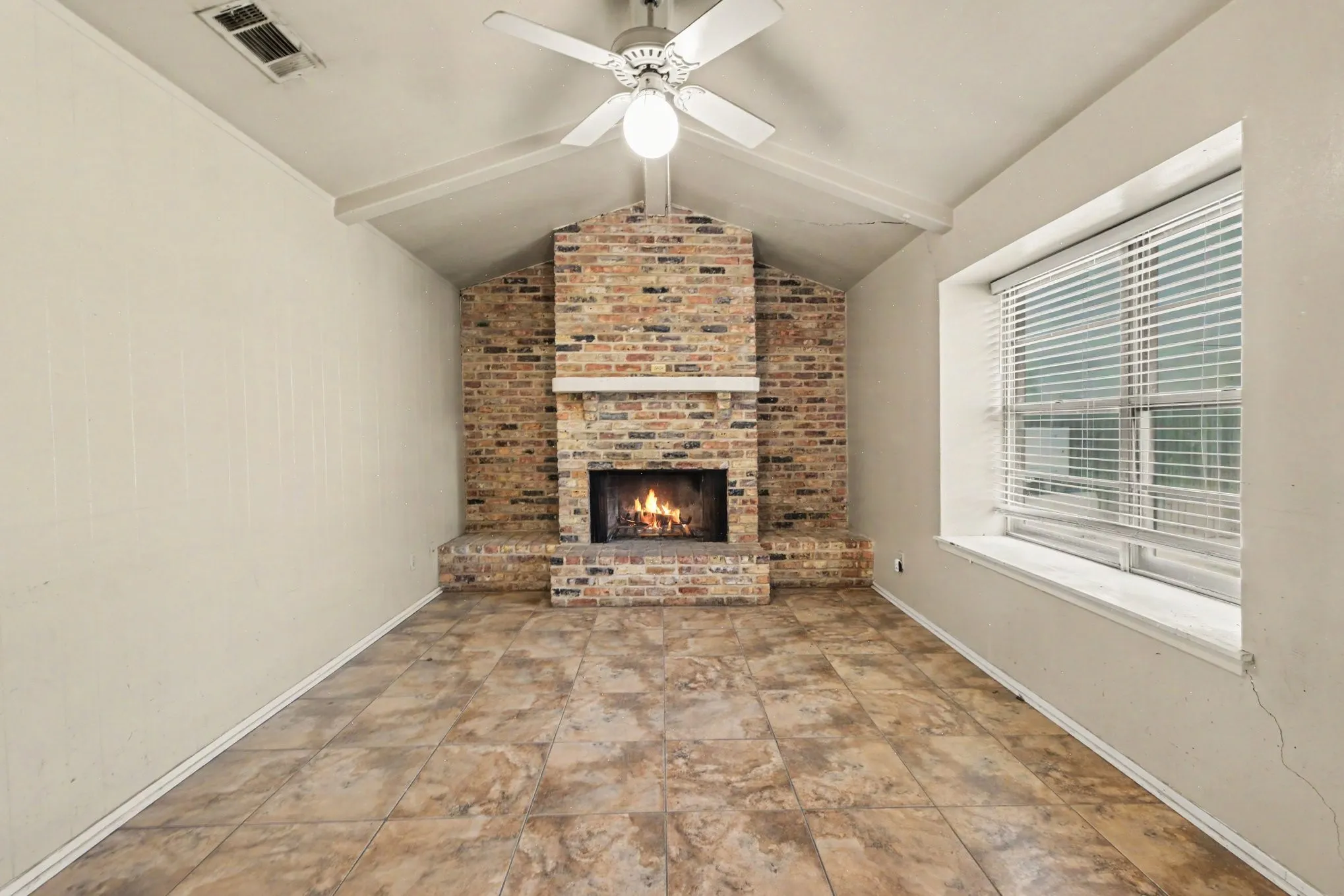 Vaulted ceiling with exposed beam, brick fireplace with mantel, window with white blinds, ceiling fan with integrated lighting, and neutral tone tile flooring