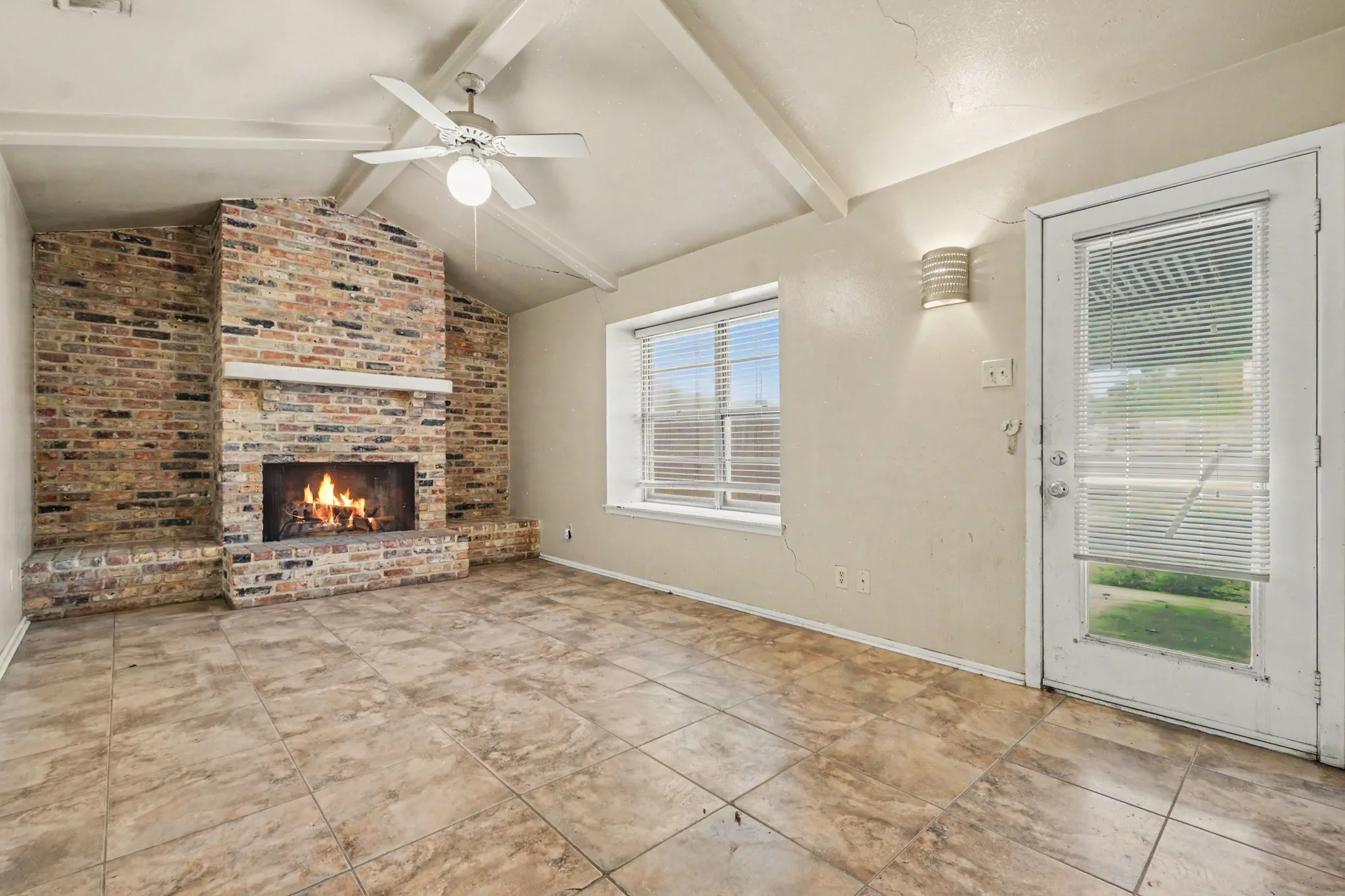 Vaulted ceiling with exposed beams, brick-clad fireplace with mantel, tiled flooring, ceiling fan, and access to exterior via a glass-paneled door