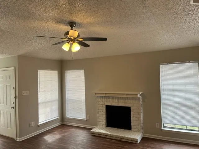 Wood-finish flooring, brick fireplace with mantel, ceiling fan with light fixture, multiple windows with blinds, and a paneled entry door