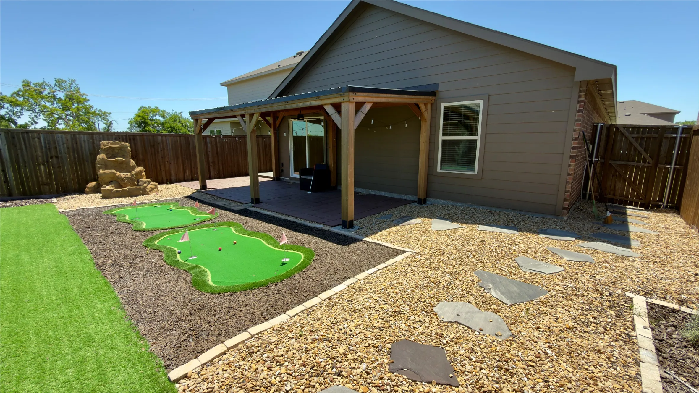 Backyard featuring a covered patio with string lighting, two putting greens, a water feature, and a stone walkway