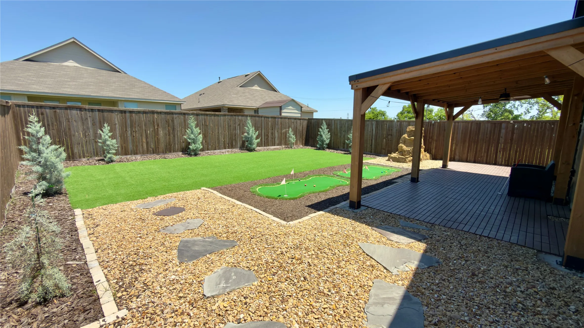 Expansive backyard featuring a covered patio with wood beam construction, a putting green, artificial turf lawn, and a pebble stone pathway with stepping stones