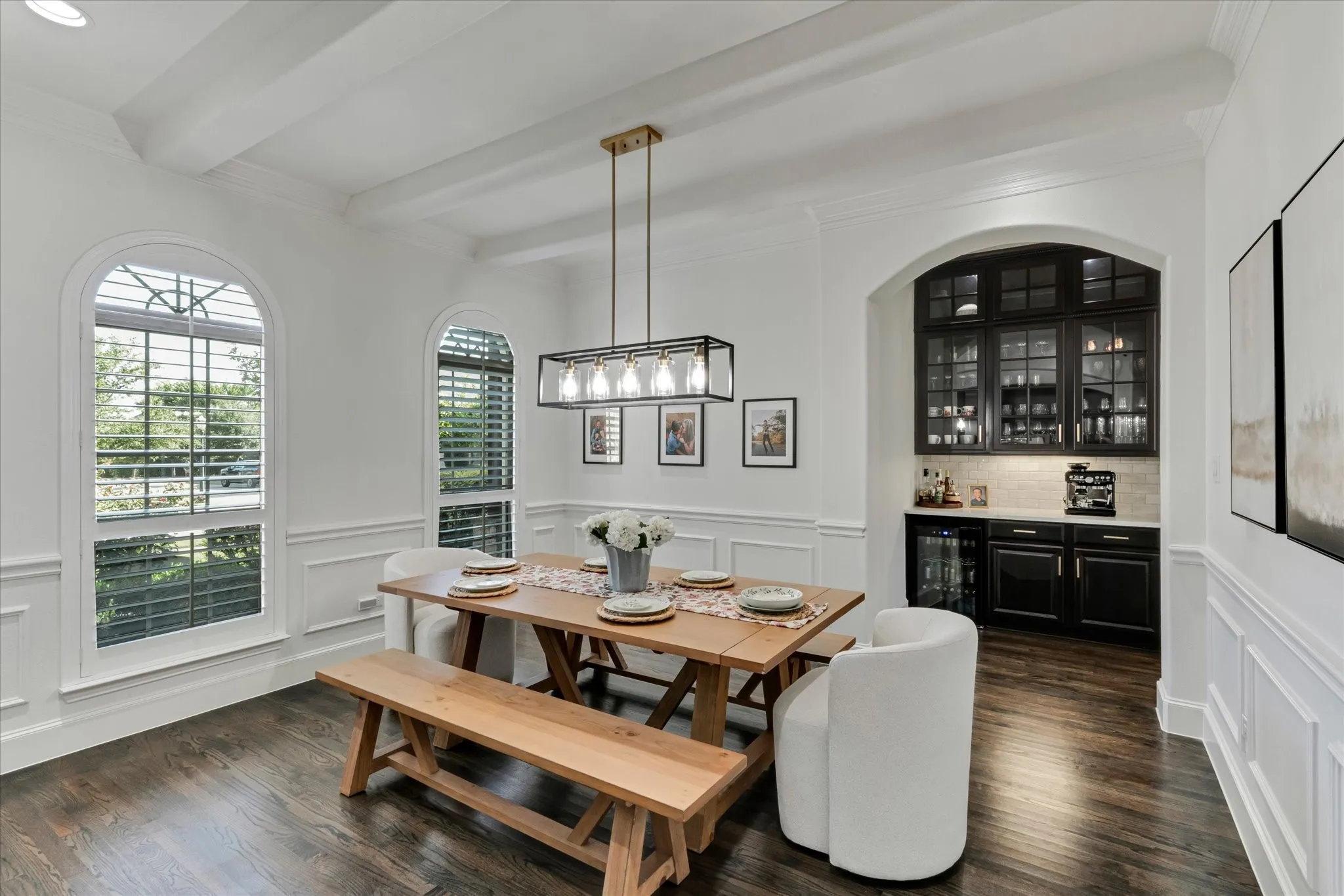 Dark wood-finish flooring, white wainscoting, arched windows with plantation shutters, a coffered ceiling, and a built-in wet bar with dark cabinetry