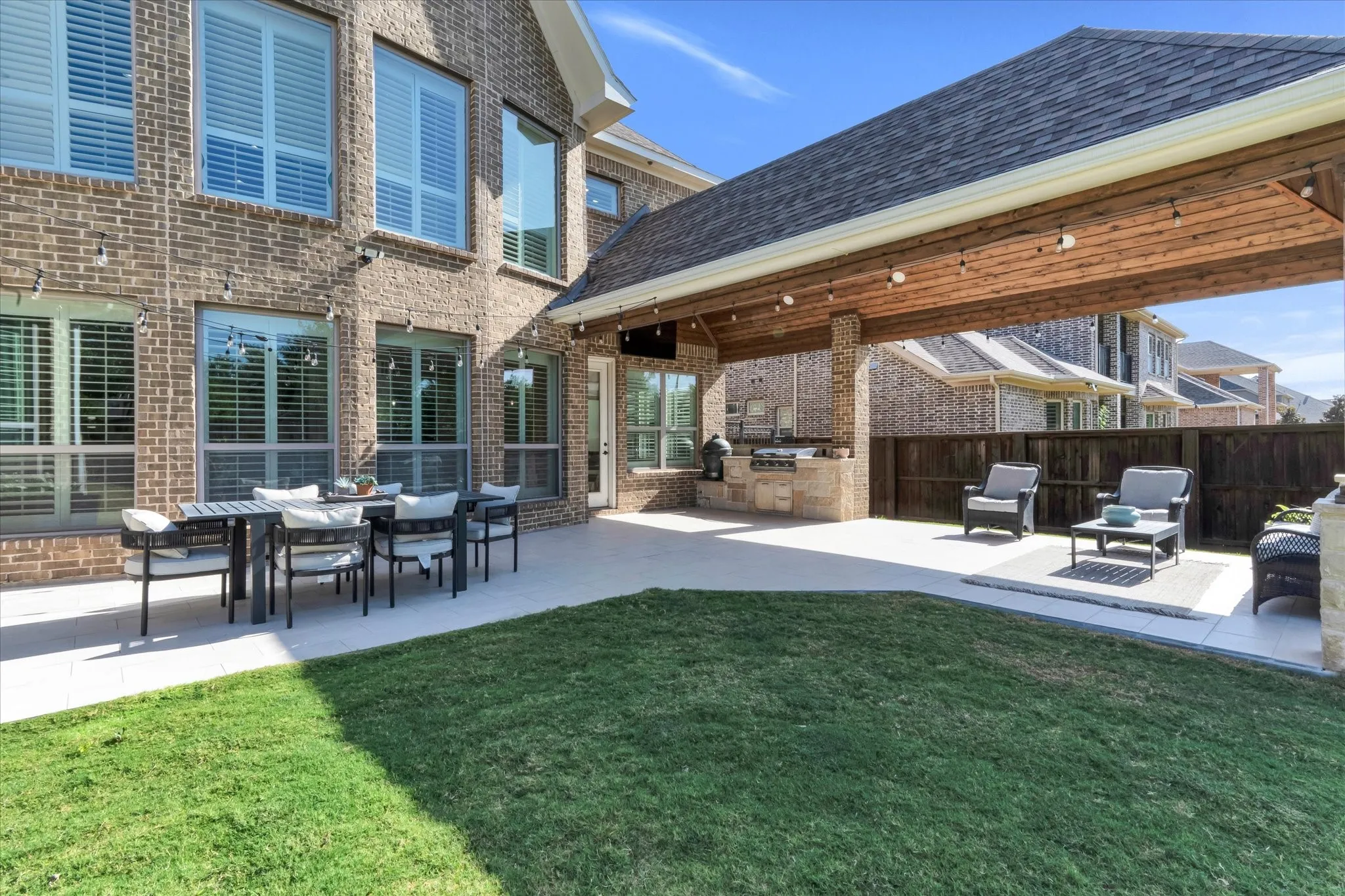 Brick exterior with multiple windows featuring plantation shutters, covered patio with wood-paneled ceiling, built-in outdoor kitchen, and concrete patio