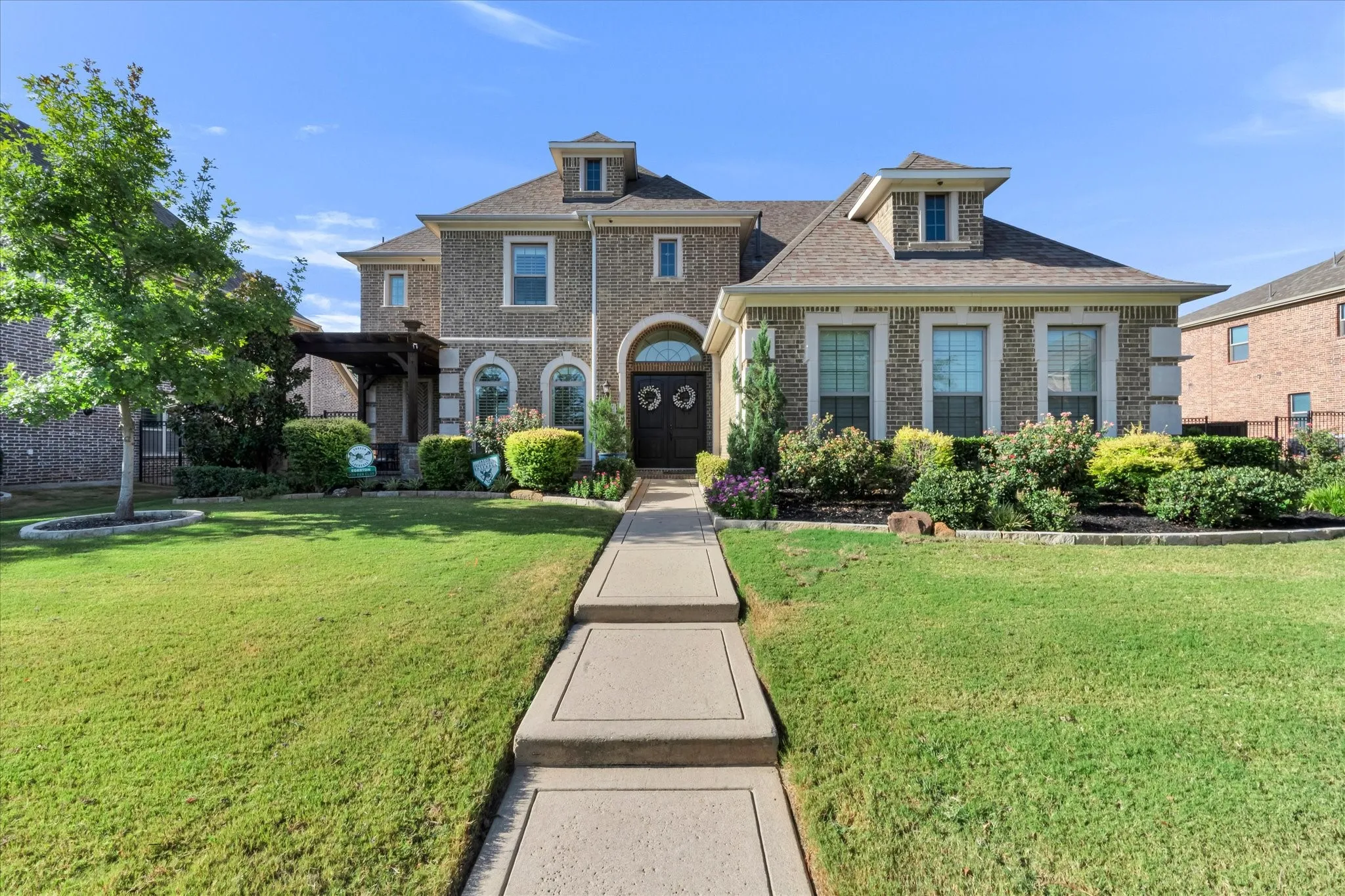 Brick facade with dormer windows, arched double entry doors, front porch pergola, and a concrete pathway