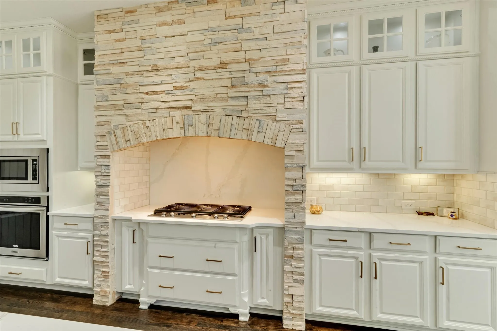Stacked stone range hood with an arched opening, stainless steel gas cooktop, white cabinetry with glass-front uppers, and subway tile backsplash