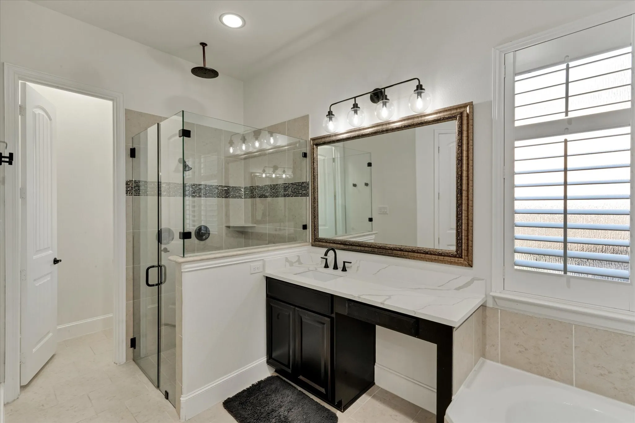 Glass-enclosed shower with a rainfall showerhead, vanity with a white countertop and dark cabinetry, large framed mirror, and plantation shutters