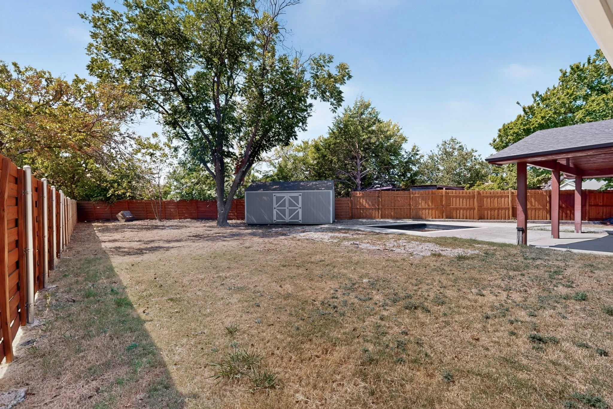 Expansive backyard with a wood privacy fence, mature trees, a storage shed, and a covered patio
