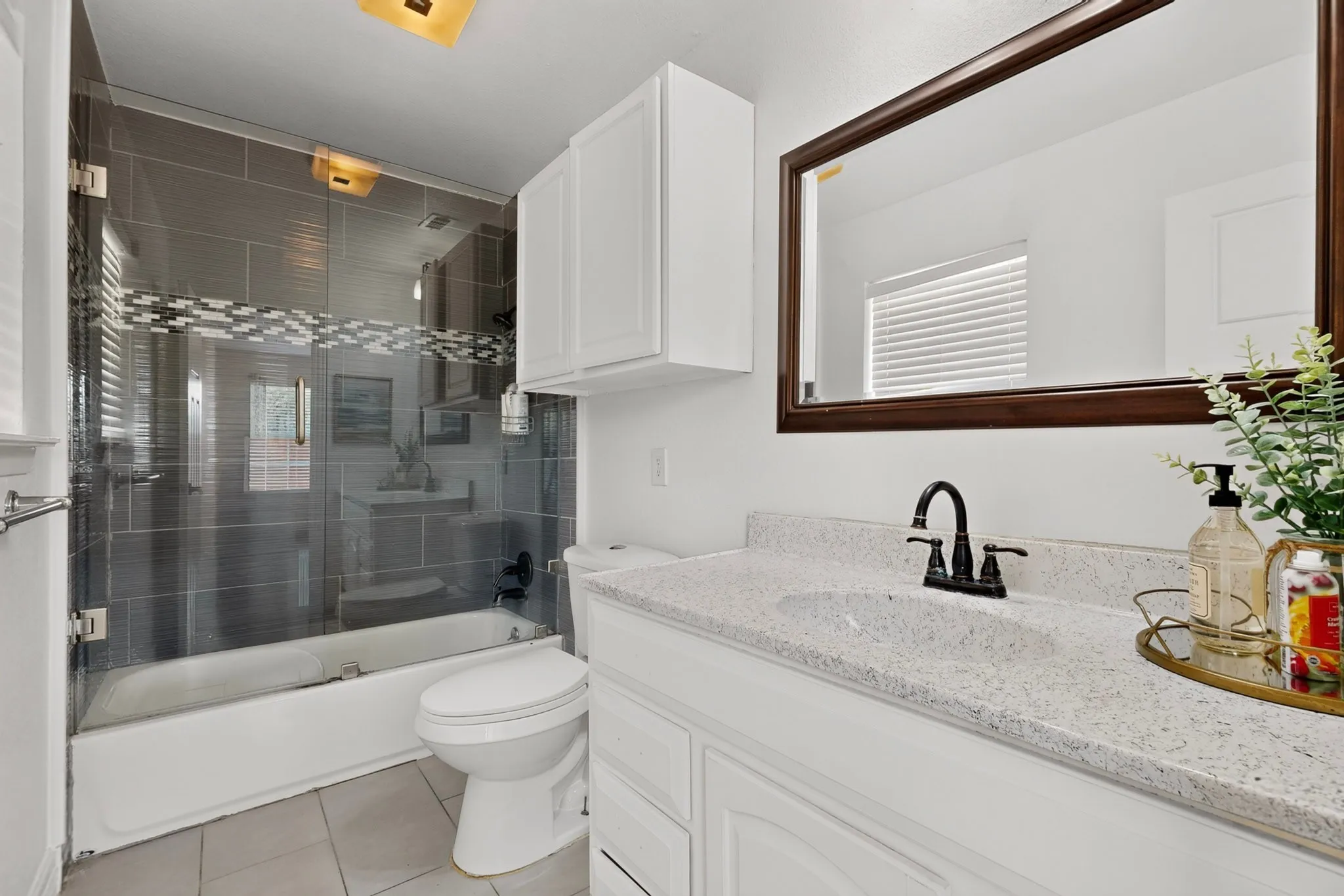 Glass-enclosed tub and shower with gray tile surround, white vanity with integrated sink, speckled countertop, dark-framed mirror, and white cabinetry