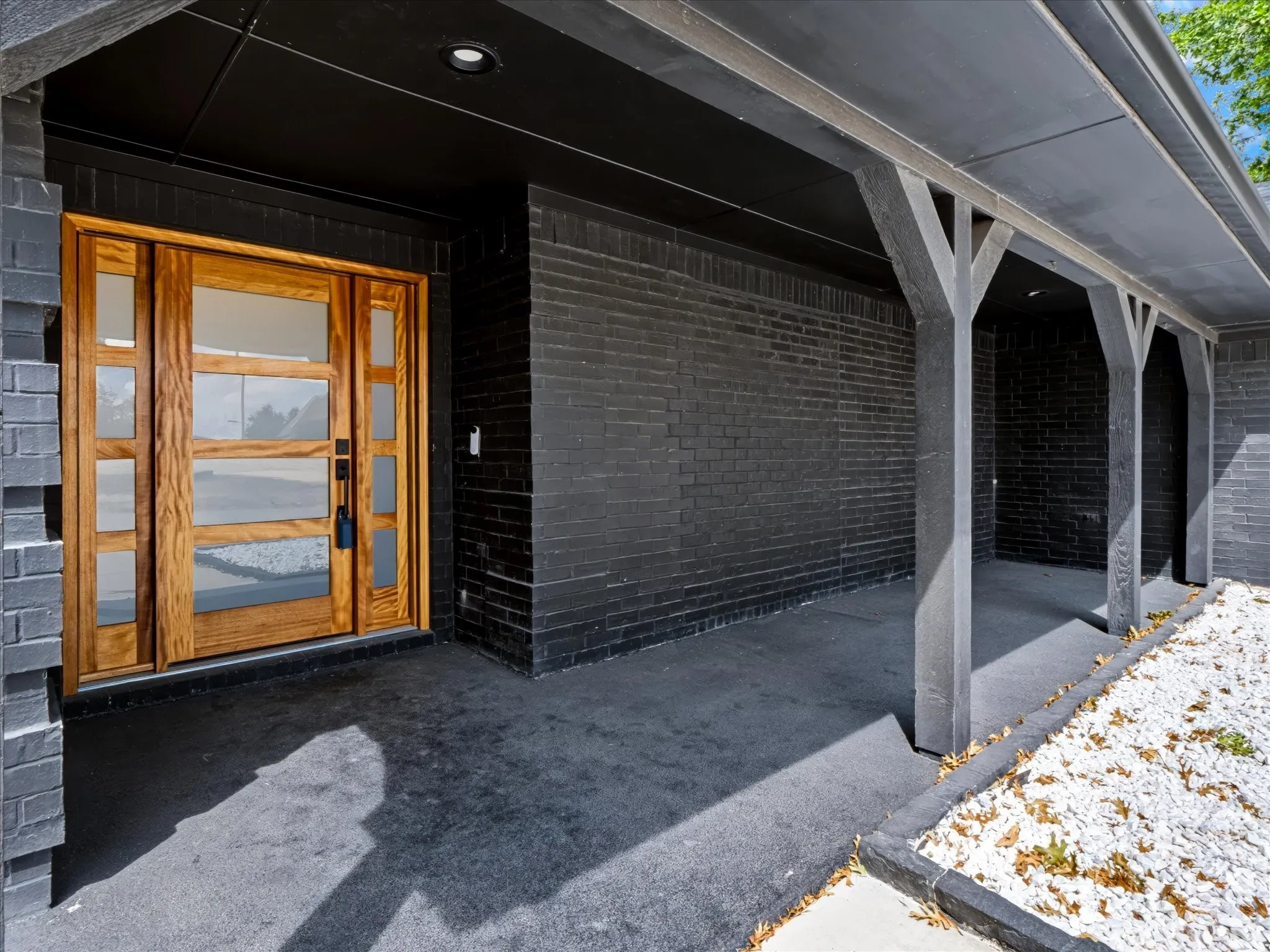 Natural wood front door with frosted glass panels, black brick exterior, covered entry, black soffit with recessed lighting, and white decorative rock landscaping