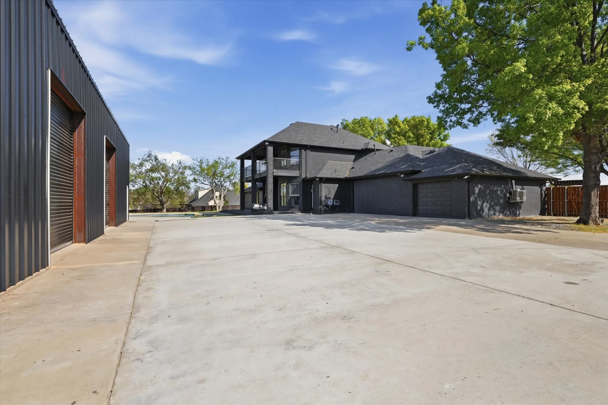 Expansive concrete driveway, corrugated metal building with two roll-up doors, multi-story structure with balconies, and a three-car garage