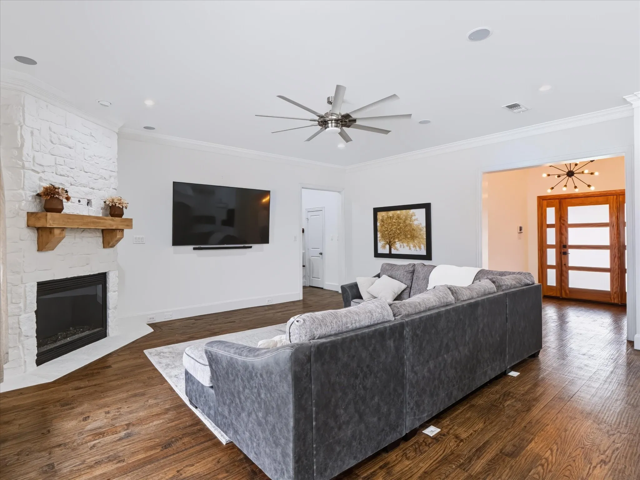 Wood-finish flooring throughout, white stone-clad fireplace with a wood mantel, crown molding, modern ceiling fan, and recessed lighting