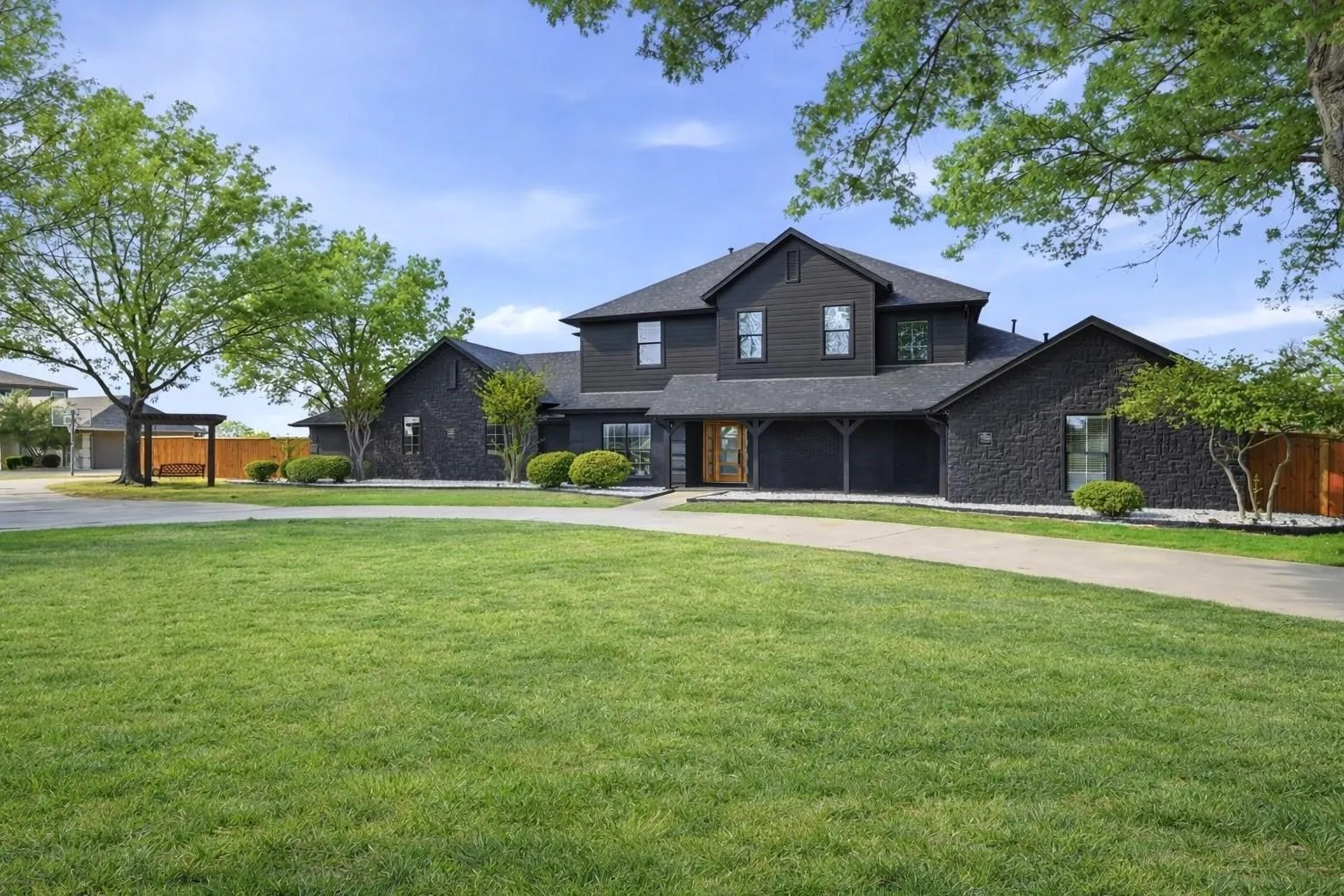 Expansive front lawn with a paved driveway, featuring a two-story residence with dark siding, a covered entry, and a side pergola