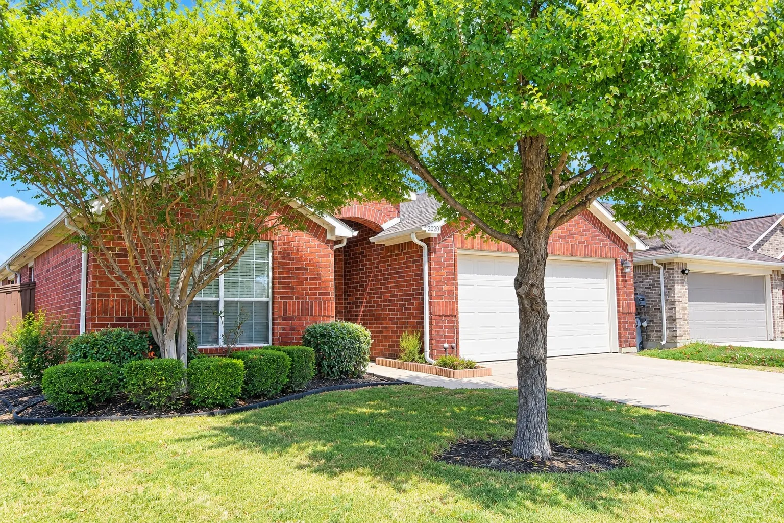 Brick facade residence with a two-car garage, manicured lawn, and mature trees