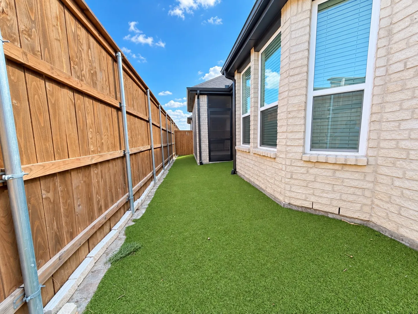 Fenced exterior corridor featuring artificial turf, light brick facade, and multiple windows with blinds