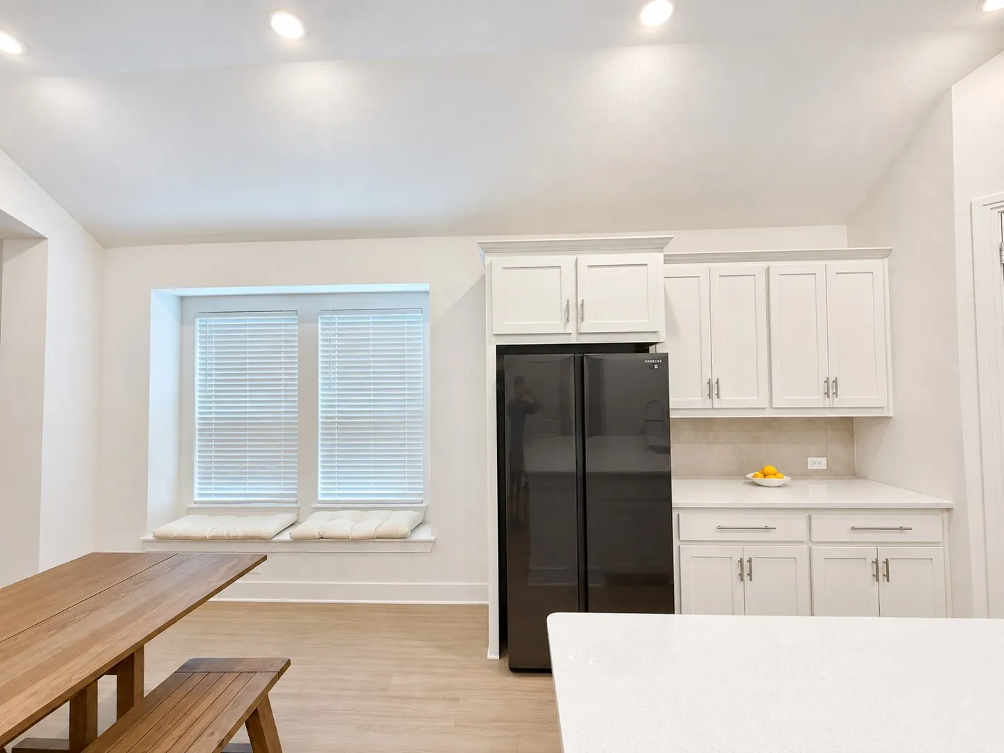 Recessed lighting, white shaker-style cabinetry, light-toned wood-finish flooring, built-in window seating, and a large black refrigerator