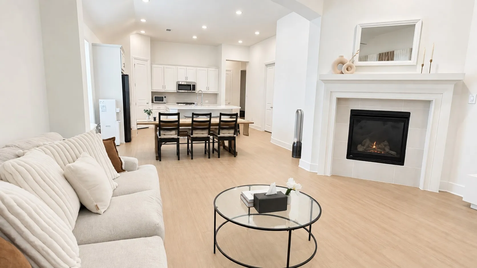 Open-concept living area featuring light wood-finish flooring, a built-in fireplace with a mantel, recessed lighting, and a kitchen with white cabinetry and stainless steel appliances