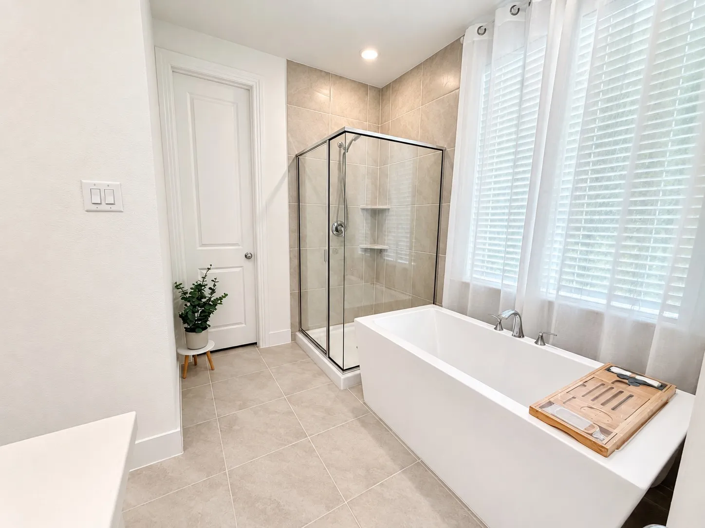 Freestanding soaking tub with chrome fixtures, tiled walk-in shower with glass enclosure, large windows with white blinds, and light-toned floor tile
