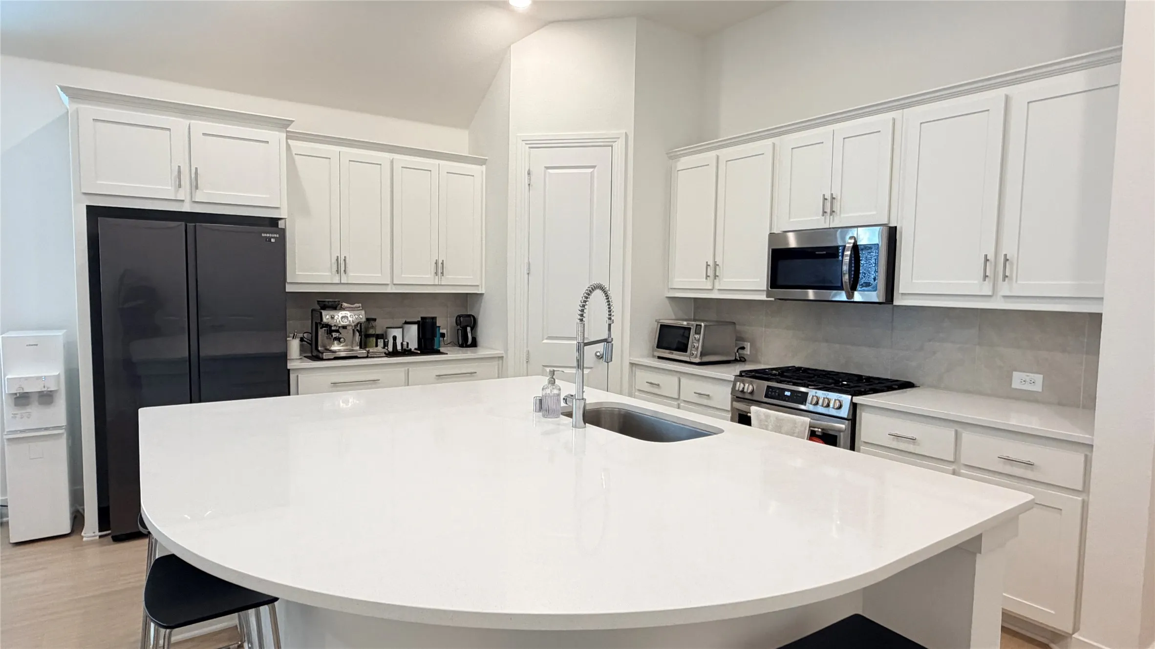 Expansive kitchen island with a curved countertop, stainless steel appliances, white cabinetry with silver-tone hardware, and a tile backsplash