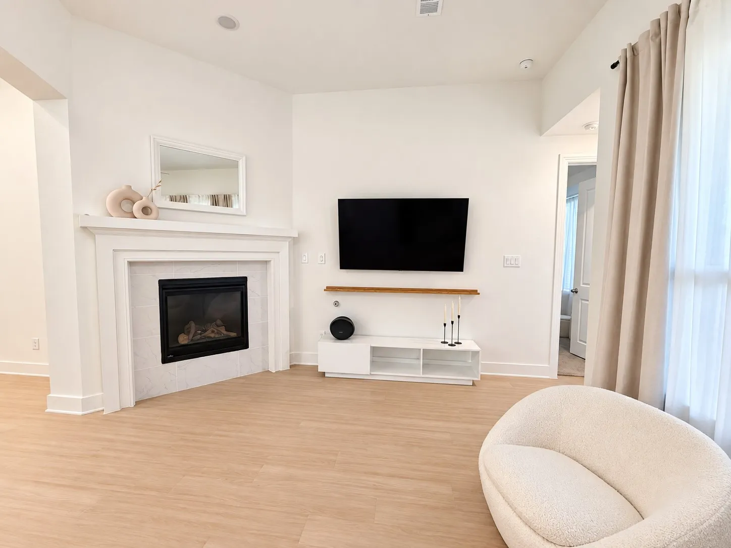 Corner fireplace with white mantel and surround, light wood-finish flooring, recessed lighting, and a large window with curtain rod