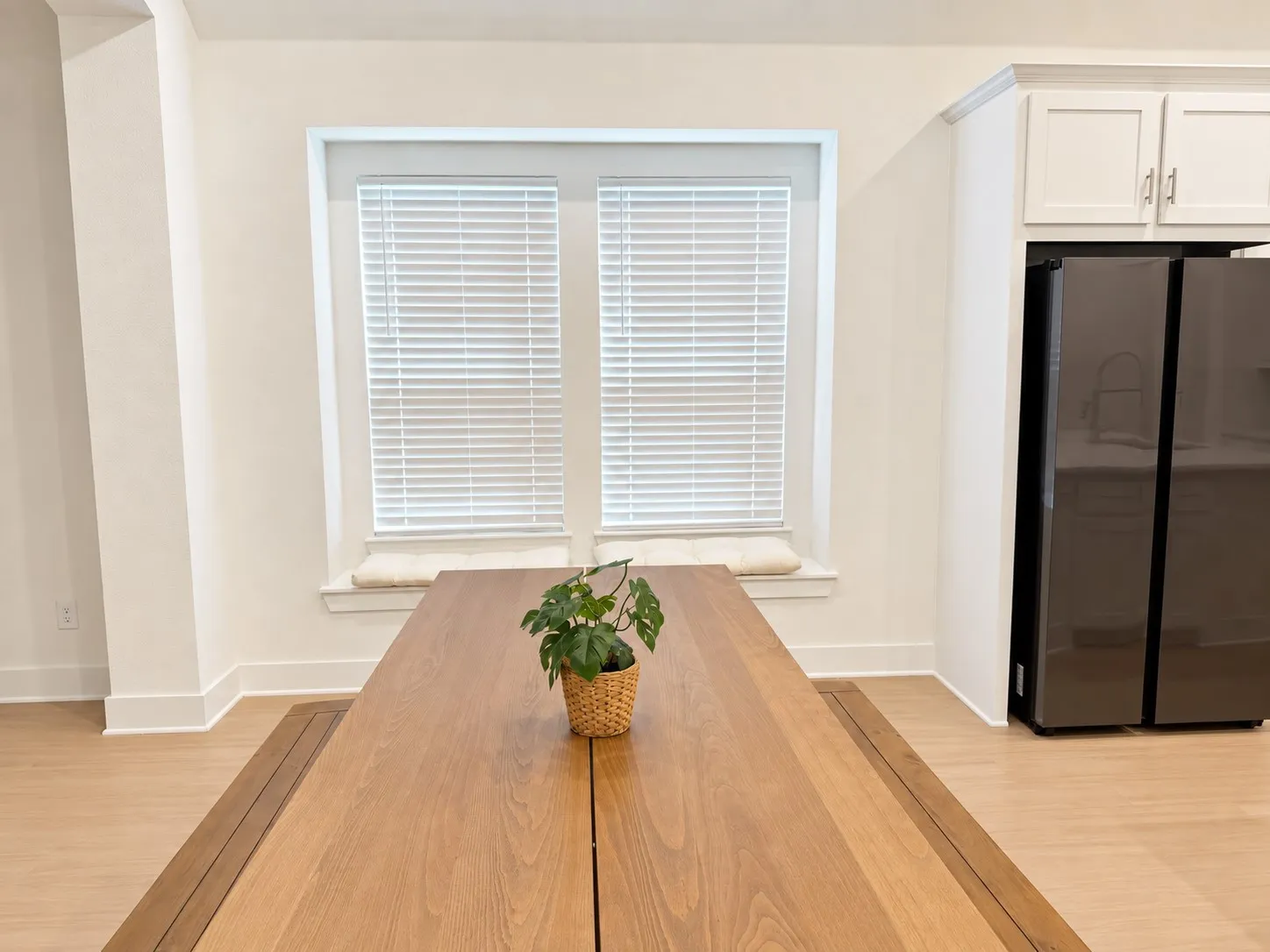 Light wood-finish flooring, built-in window seat, dual windows with blinds, white upper cabinetry, and a dark stainless steel refrigerator