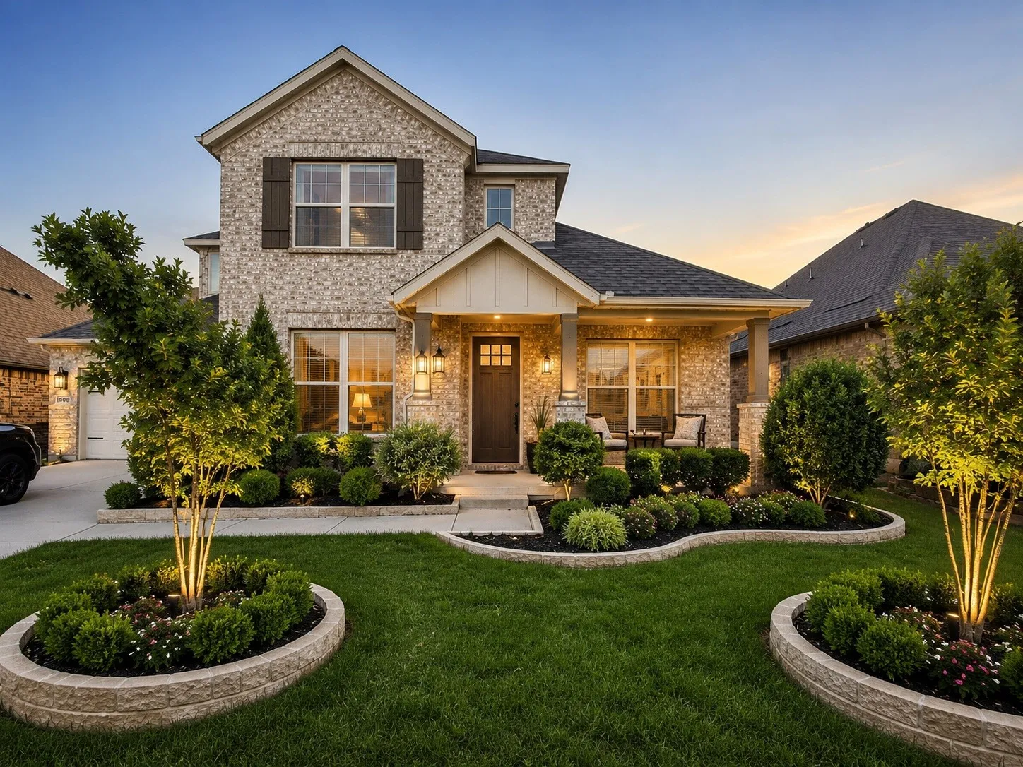 Brick exterior, dark shingle roof, covered front porch with columns, landscaped front yard with layered plantings, and concrete driveway