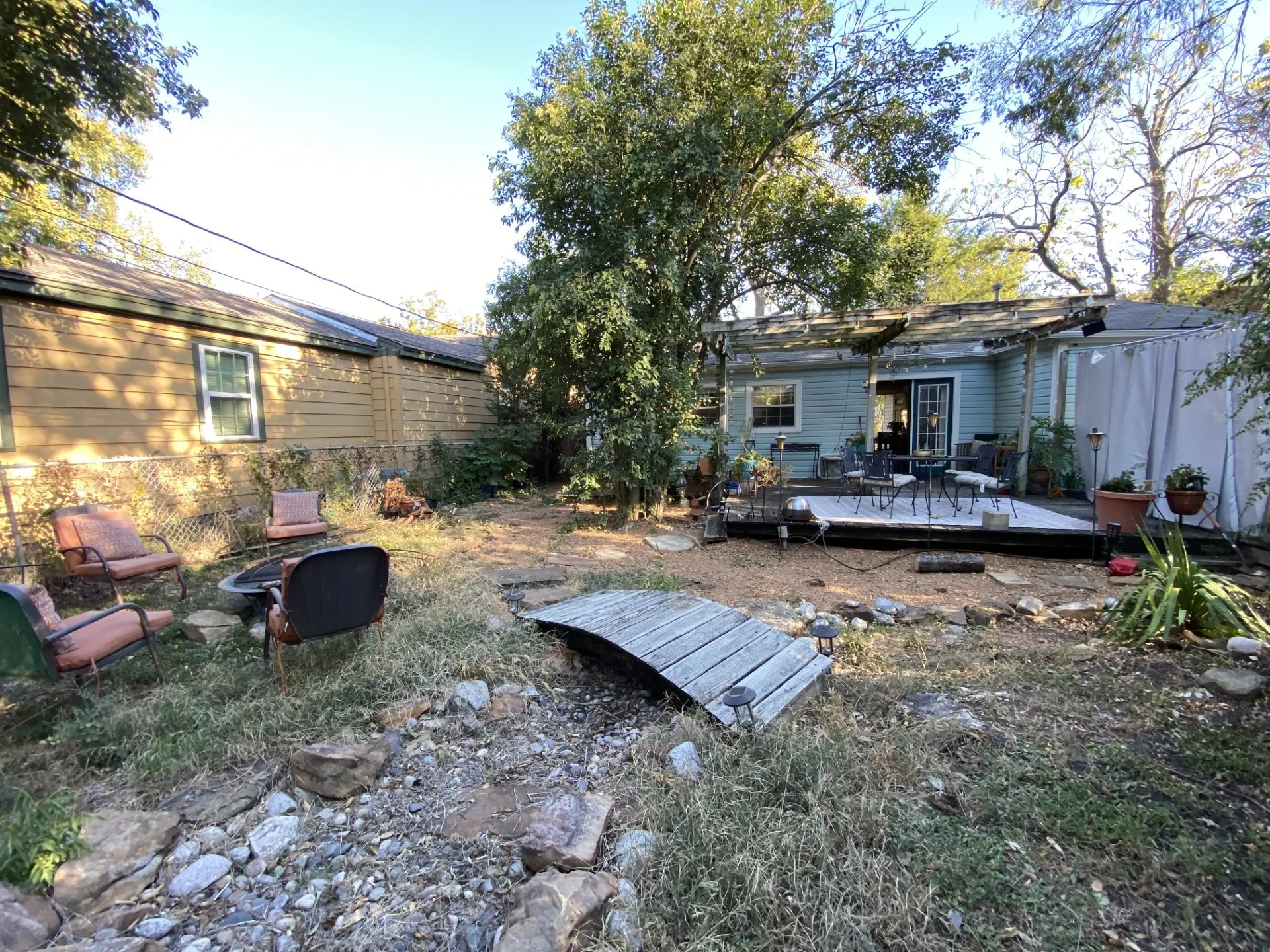 Expansive backyard featuring a raised wooden deck, a covered patio area, and a decorative wooden bridge over a dry creek bed