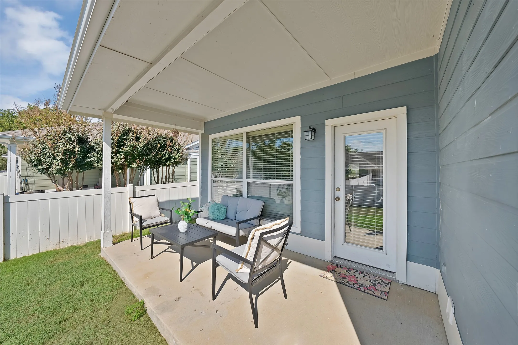 Covered concrete patio with a white privacy fence, blue siding, a glass-paned door, and an exterior wall light