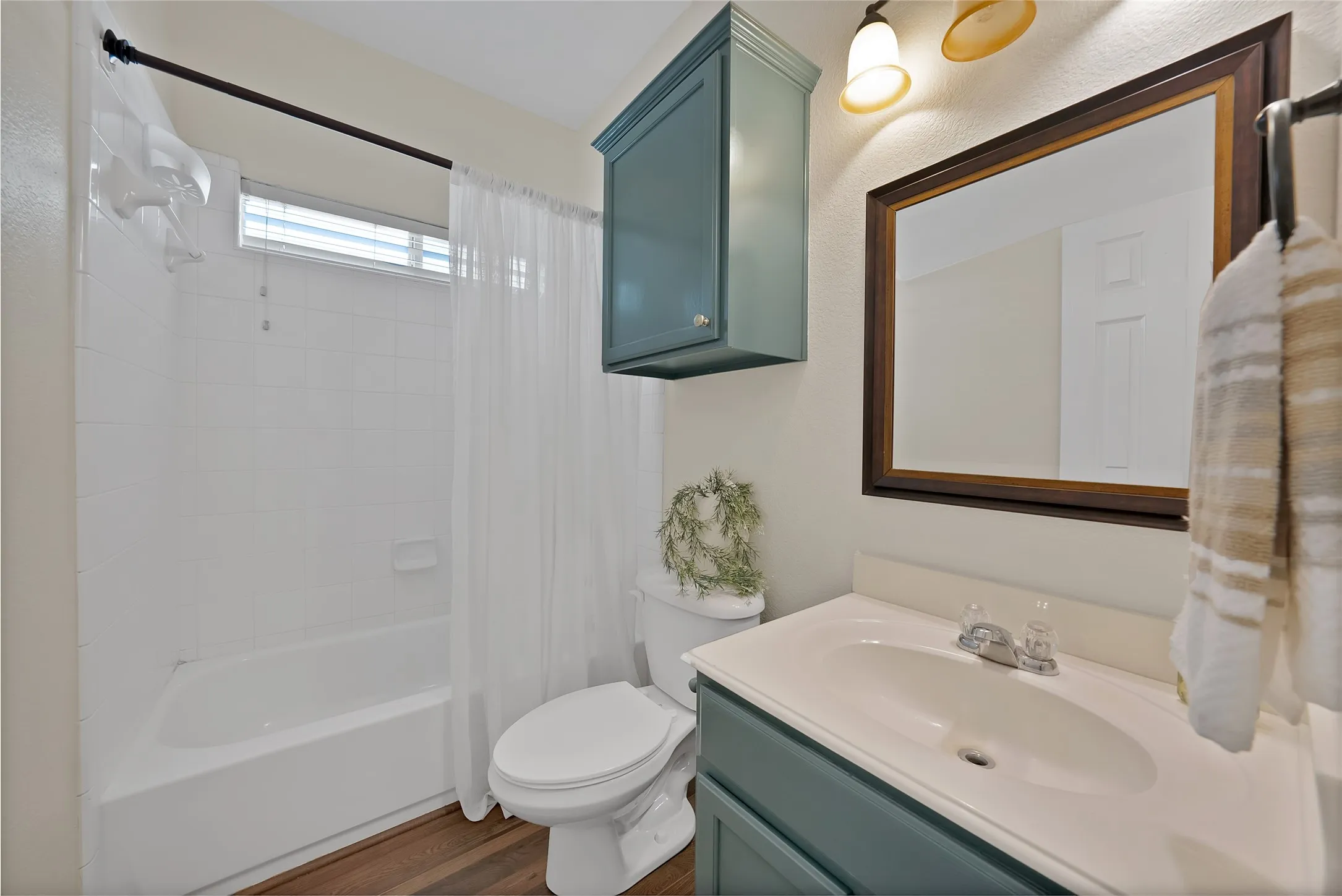 Bathroom featuring a white bathtub with tile surround, wood-finish flooring, a toilet, a vanity with a white countertop and teal cabinetry, and a framed mirror