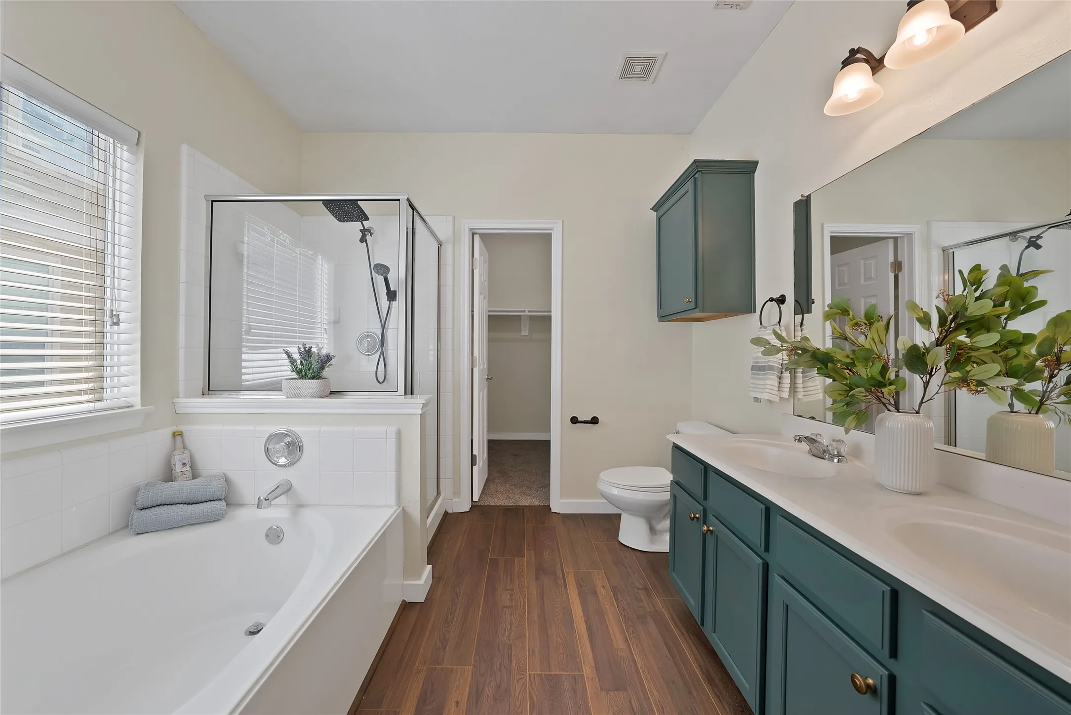 Dual vanity with white countertops, wood-finish flooring, a built-in tub, and a separate glass-enclosed shower