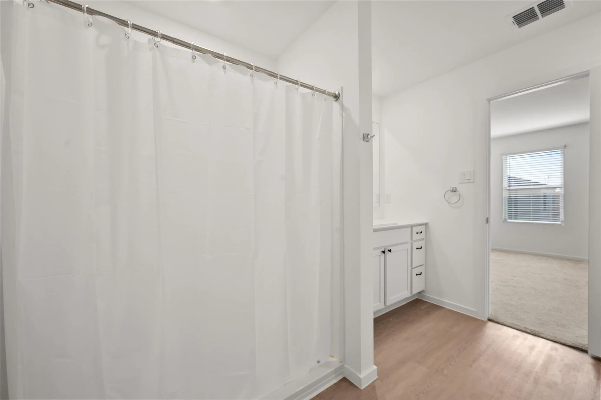 White bathroom vanity with built-in storage, light wood-finish flooring, and a shower curtain rod