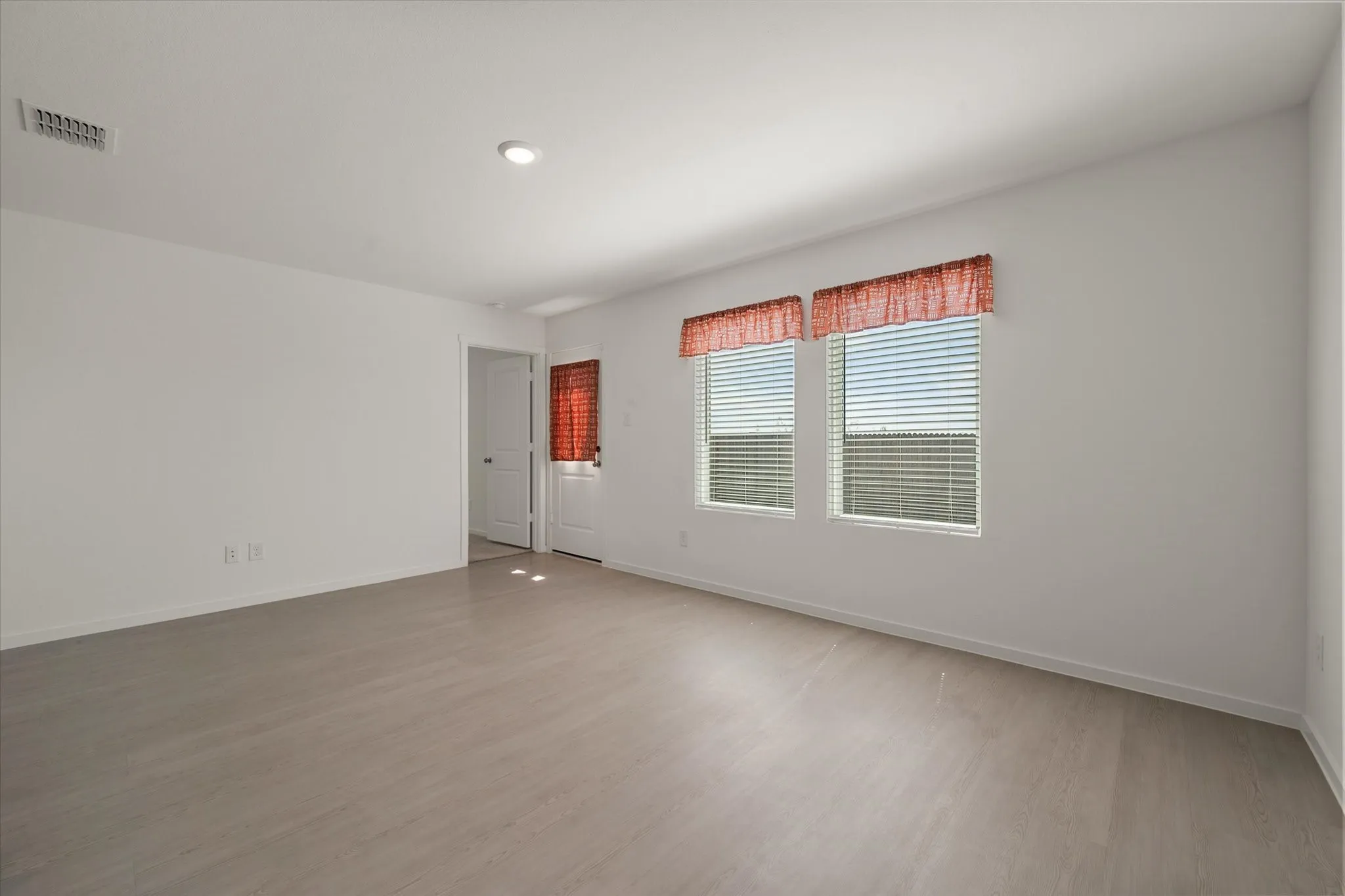 Light wood-finish flooring, two large windows with blinds, recessed lighting, white walls, and a white door with a dark handle