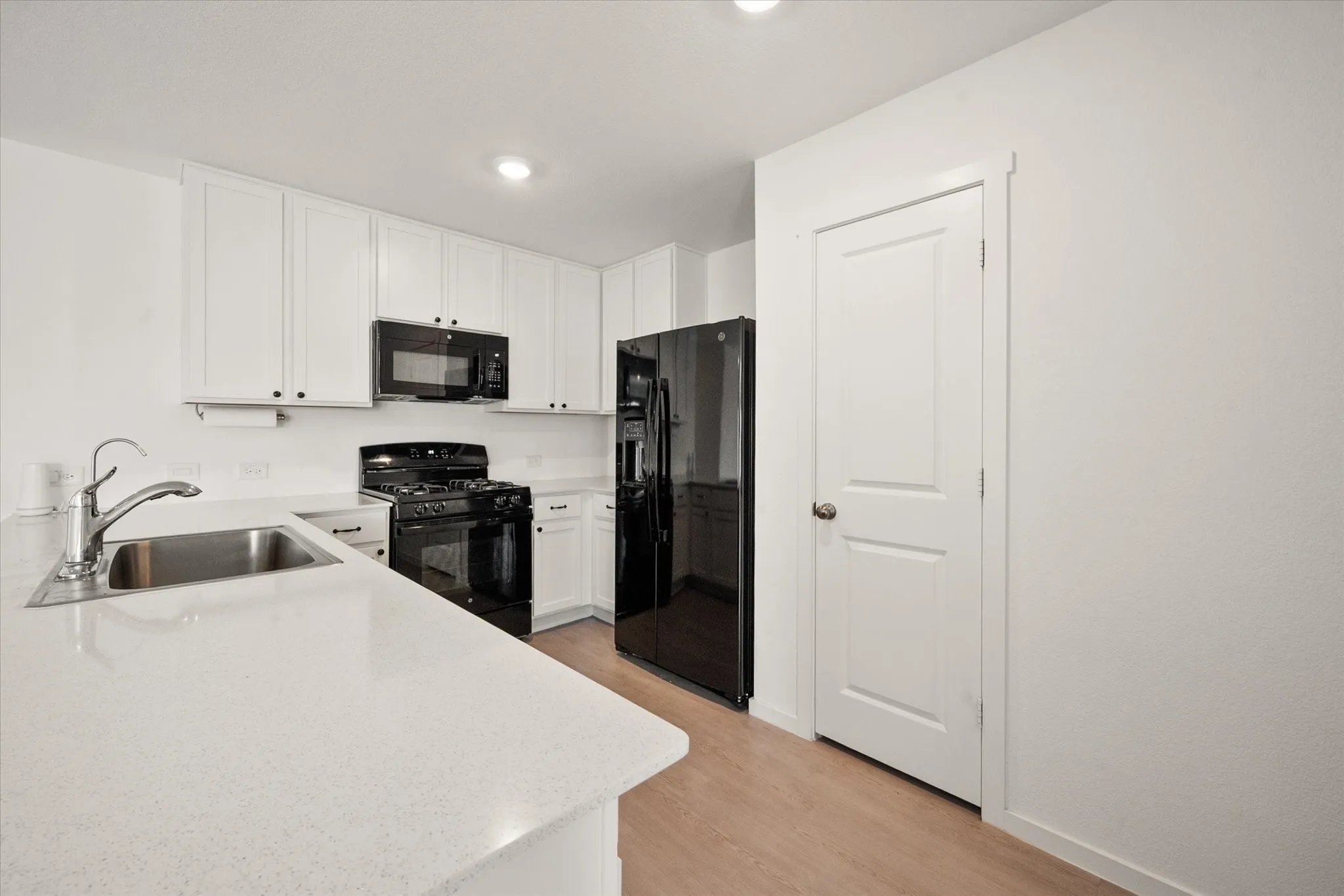 Kitchen featuring white upper and lower cabinetry, light-toned countertops, a stainless steel sink with a gooseneck faucet, black appliances, and wood-finish flooring