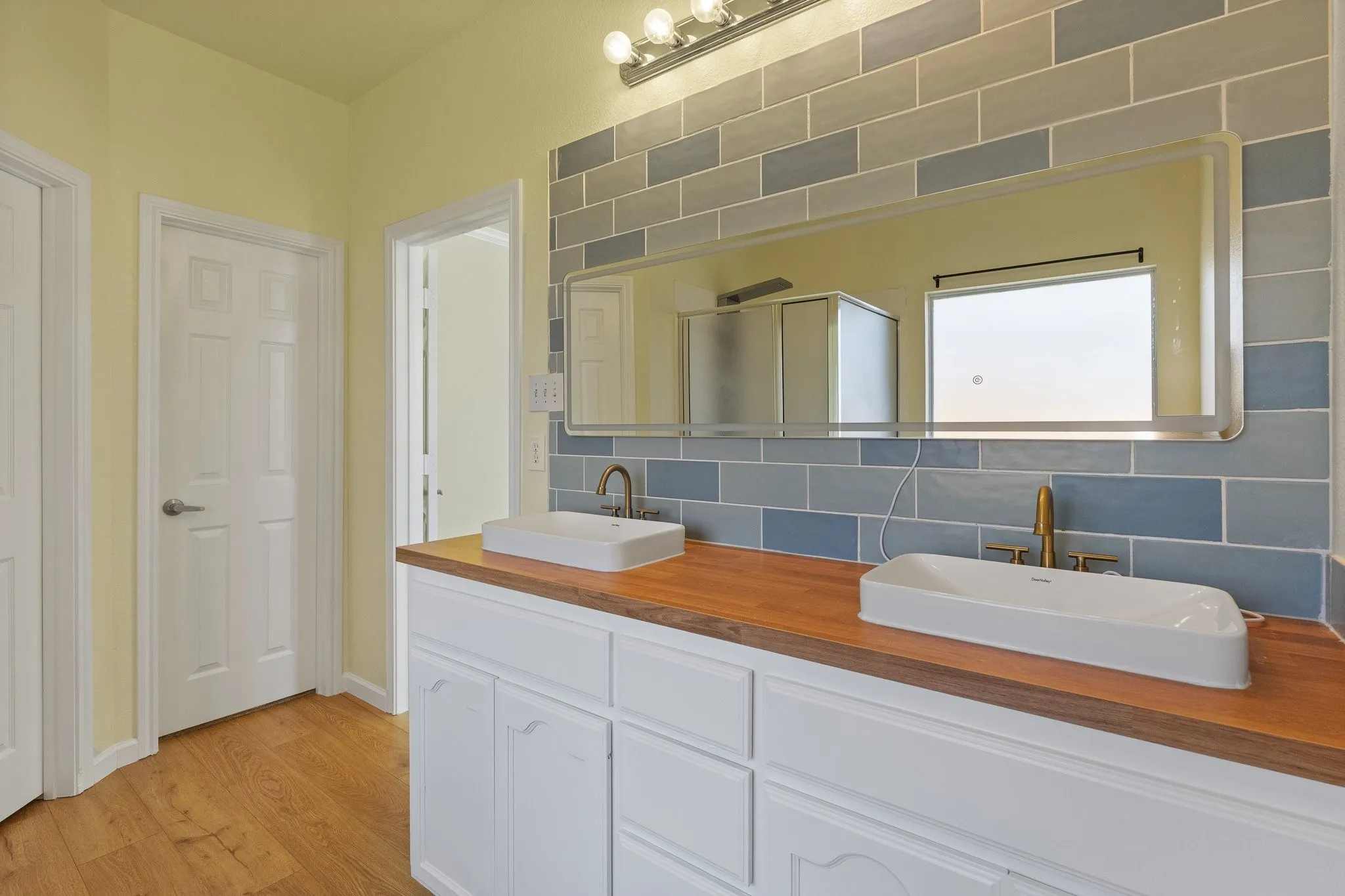 Dual vessel sinks with brass-finish faucets, wood-finish countertop, white cabinetry, and blue tile backsplash