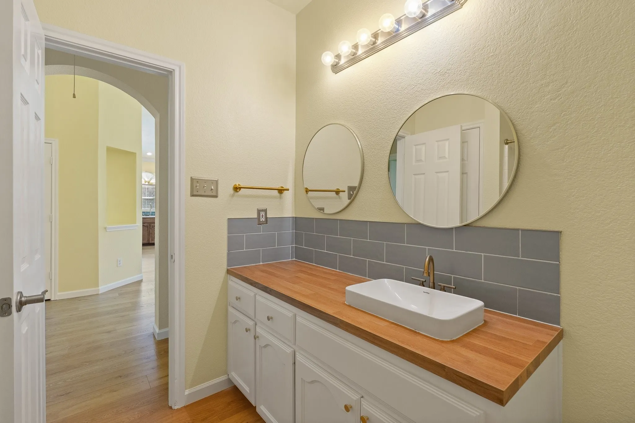 Wood-finish flooring throughout, white cabinetry with gold-tone hardware, a rectangular vessel sink, a wood-finish countertop, and gray subway tile backsplash