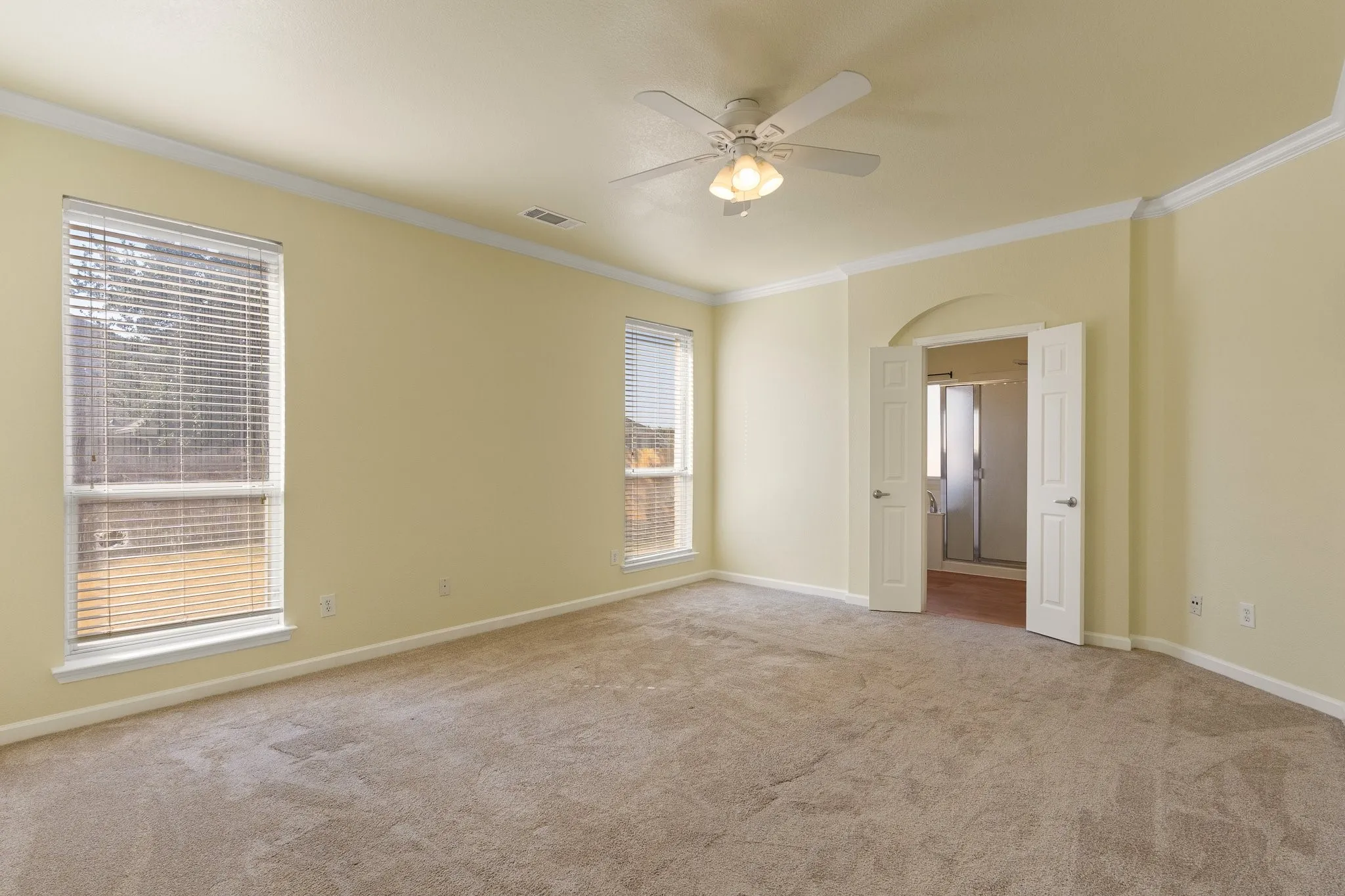 Spacious interior with neutral carpeting, two windows with blinds, a ceiling fan with integrated lighting, and an arched doorway leading to an adjacent room